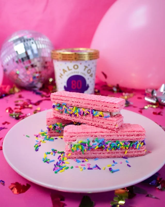 Pink layered wafers with colorful sprinkles on a white plate, decorated for a celebration with a disco ball, pink balloons, and confetti in the background.