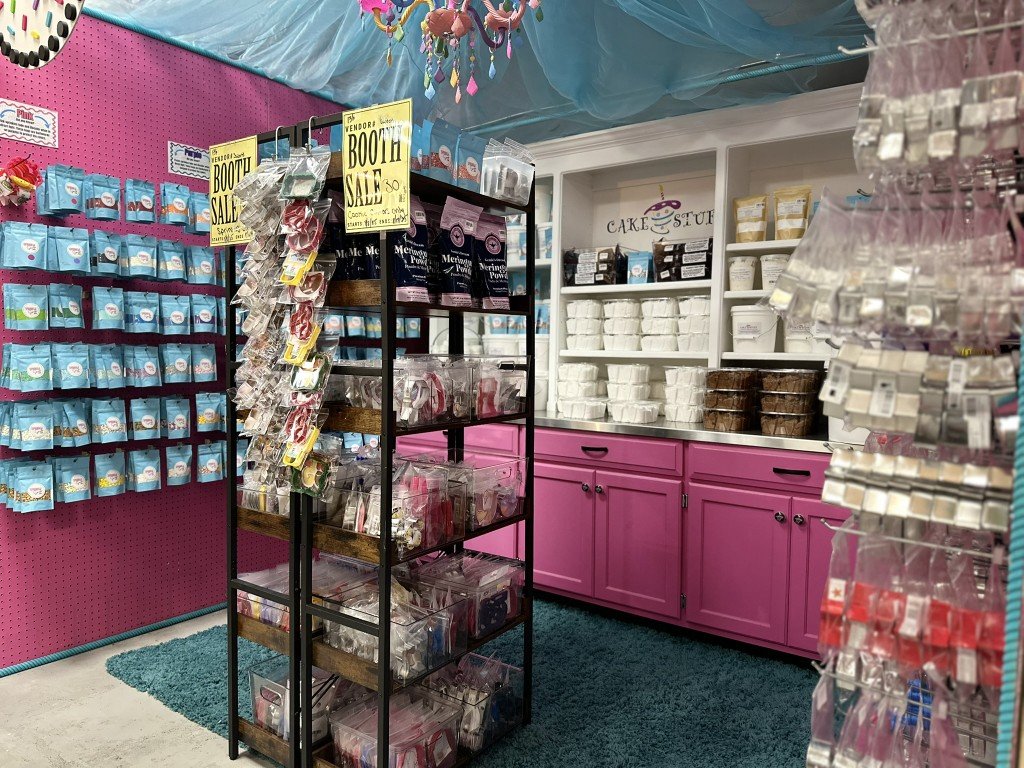 A section of a cake decorating supply store with pink cabinets, shelves stocked with cake tools and ingredients, and a pink tiling wall with hanging baking supplies.