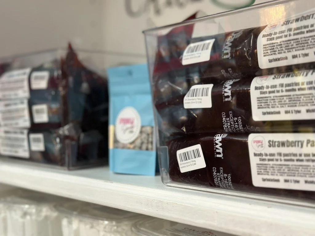 A shelf in a store with packaged food and a blue bag of sprinkles, with a focus on a clear plastic container of strawberry-flavored pastry filling with barcode labels.