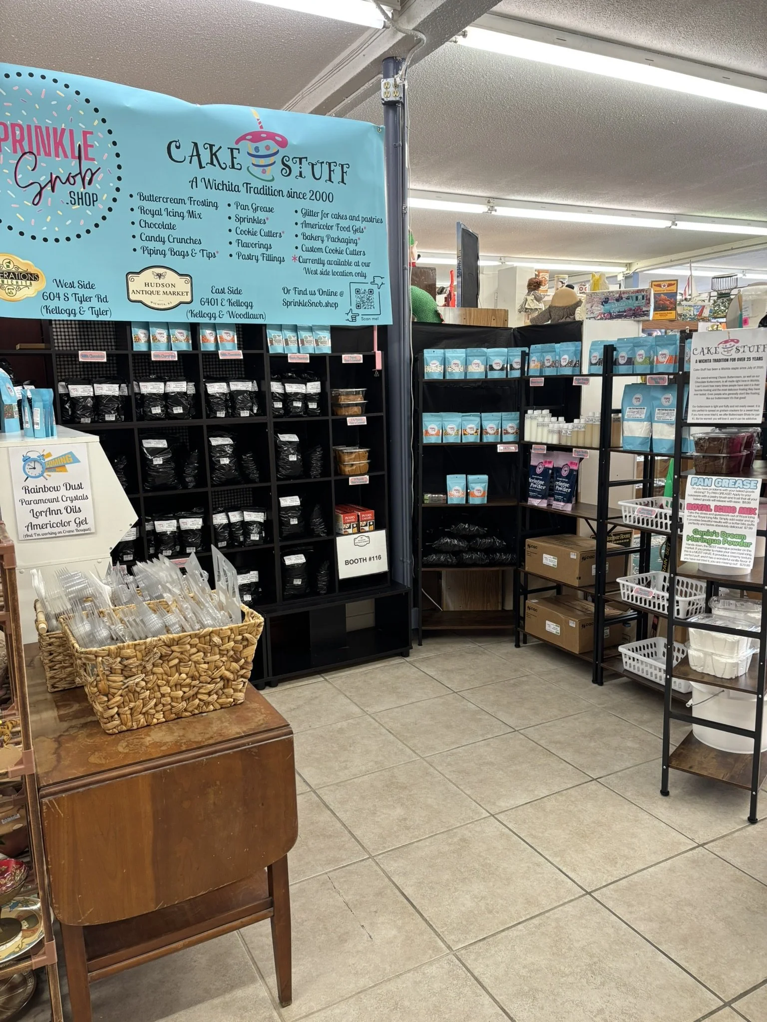 Display of cake decorating supplies at a store, including various frosting and gel products on black shelves, with signage indicating cake supplies and a basket of piping tips on a wooden table in the foreground.