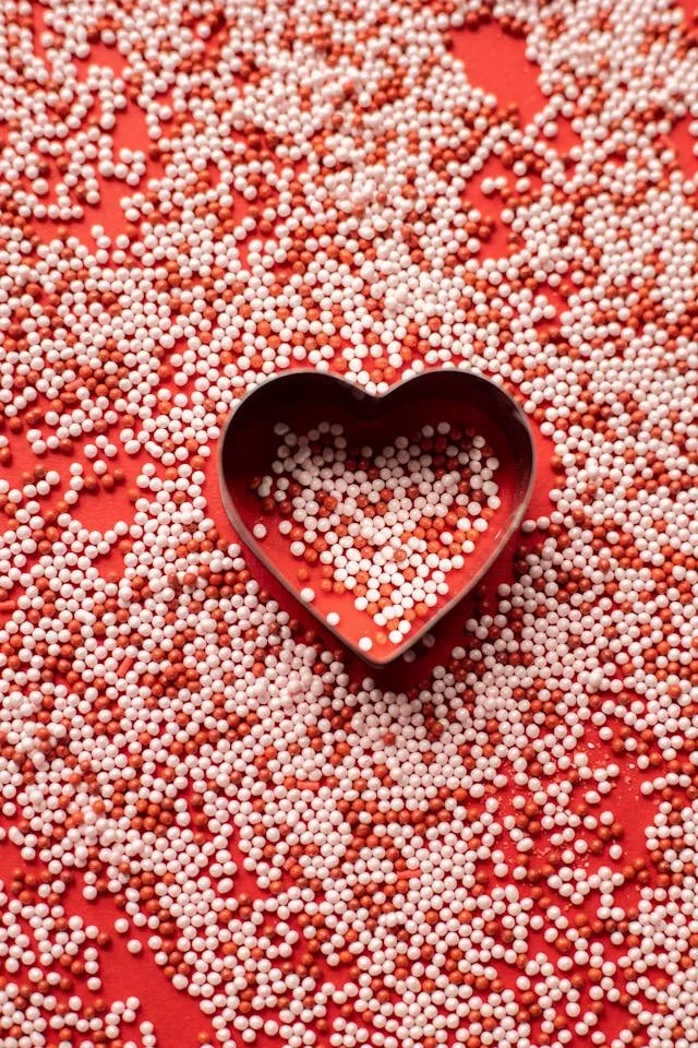 Red surface covered with numerous small white and red spherical sprinkles, with a black heart-shaped cookie cutter in the center filled with white sprinkles.