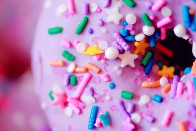 Close-up of a pink frosted donut decorated with colorful sprinkles, including stars, rods, and various small round shapes.