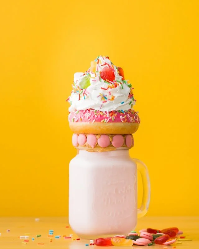 A colorful ice cream sundae topped with whipped cream, fruit, and rainbow sprinkles, served on a pink donut, placed on a mason jar filled with milk against a bright yellow background.