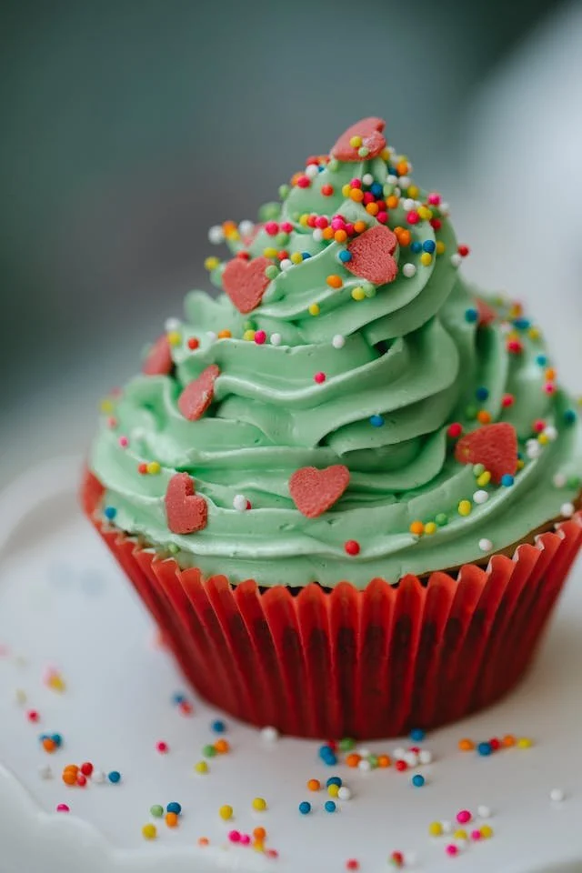 Green frosted cupcake with colorful sprinkles and pink heart-shaped decorations in a red cup on a white plate with sprinkles scattered around
