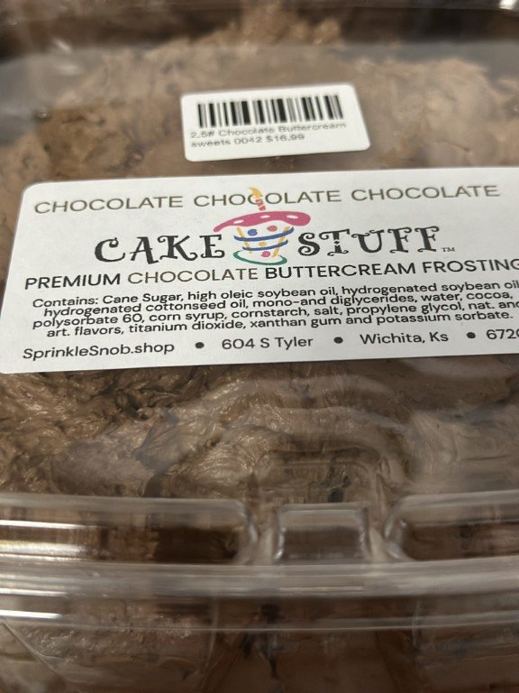 A clear plastic container of chocolate cake with a label that reads 'Cake Stuff' and ingredients including cane sugar, soybean oil, and cocoa. There is a small white sticker with a barcode and price at the top.