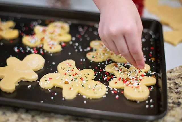 Hand decorating holiday cookies with colorful sprinkles on a baking sheet in a kitchen.