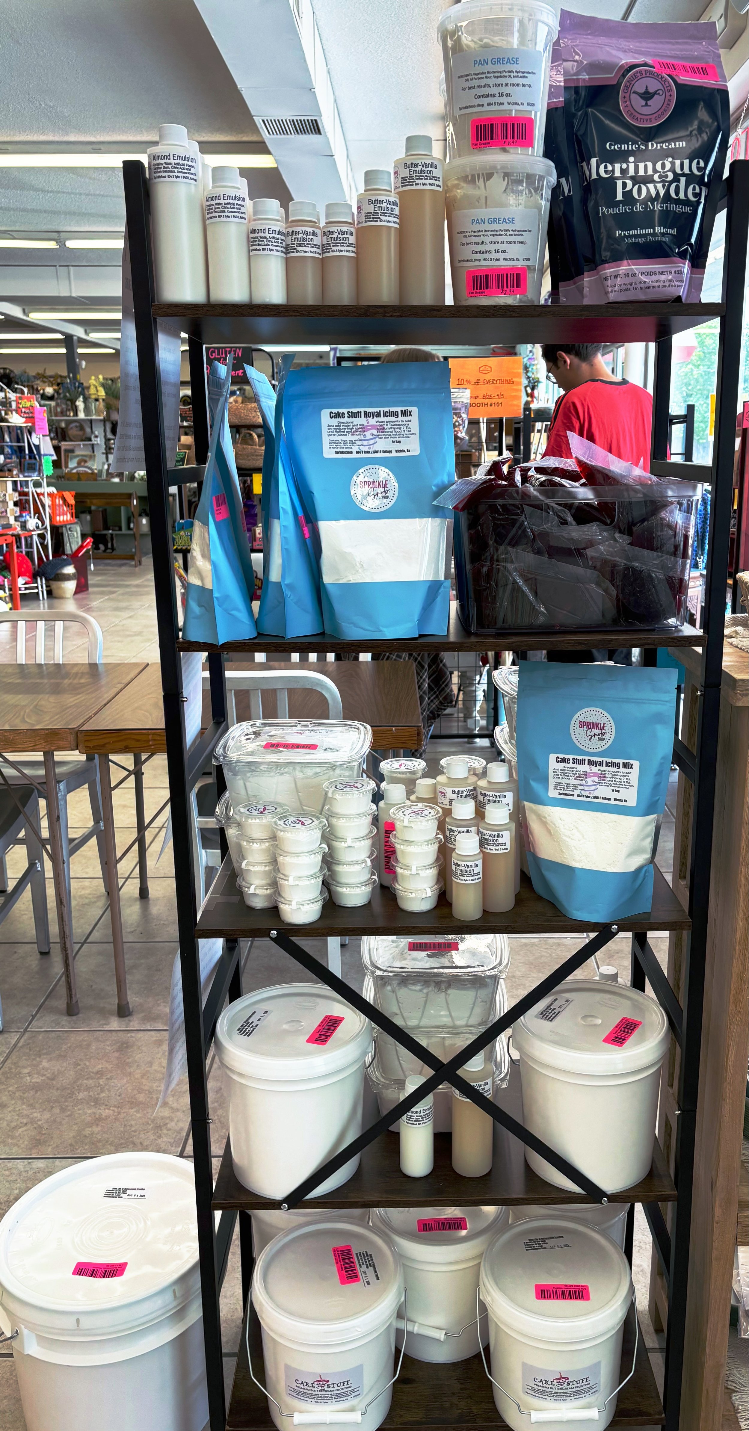 Display shelf with cake decorating supplies including butter-vanilla emulsion, almond emulsion, pan greases, meringue powder, cake mix, and various containers of icing and ingredients in a bakery or craft store.