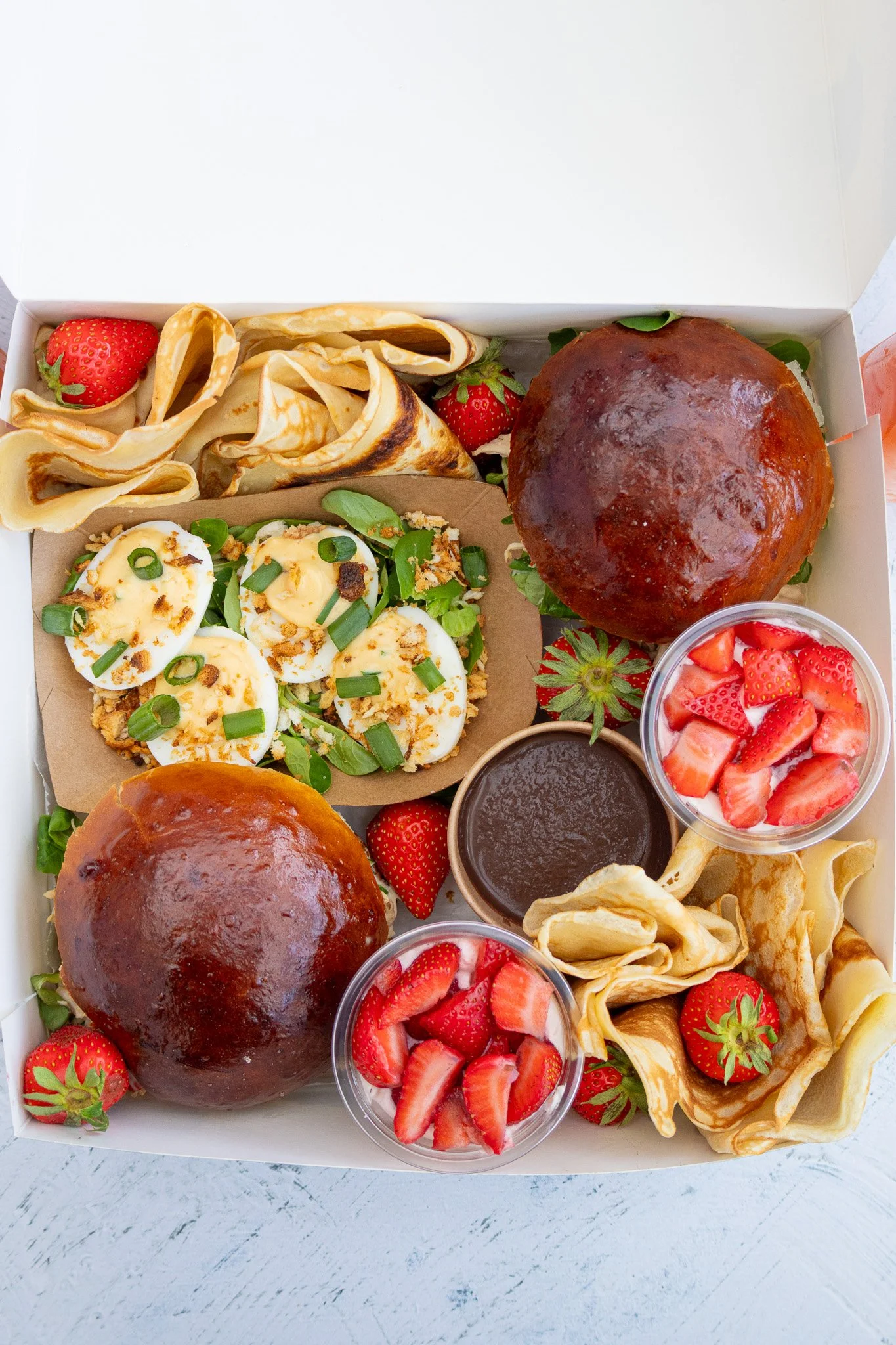 Assortiment de nourriture dans une boîte, comprenant des œufs farcis garnis de ciboulette, des fraises, des petits pains à la viande, des gaufres ou pancakes, des coupes de fraises en morceaux avec crème, deux gâteaux ou pains recouverts de glaçage o