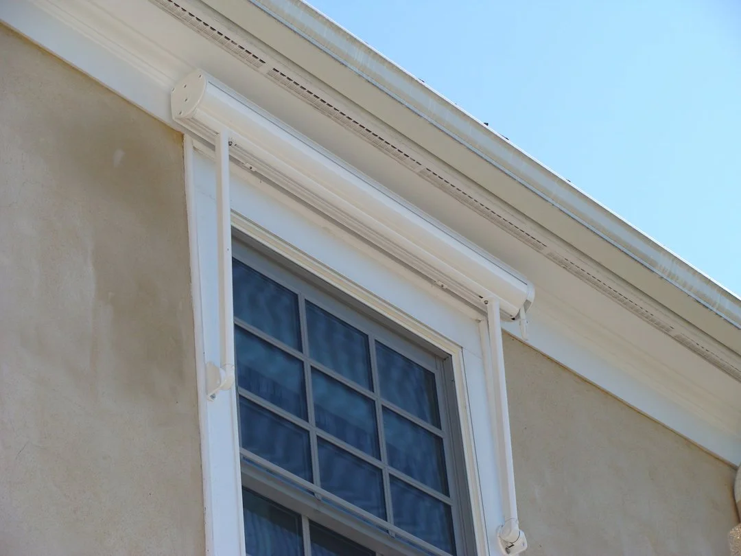 Close-up of house window with white window frame, beige stucco wall, and a white retractable awning above, under a clear blue sky.