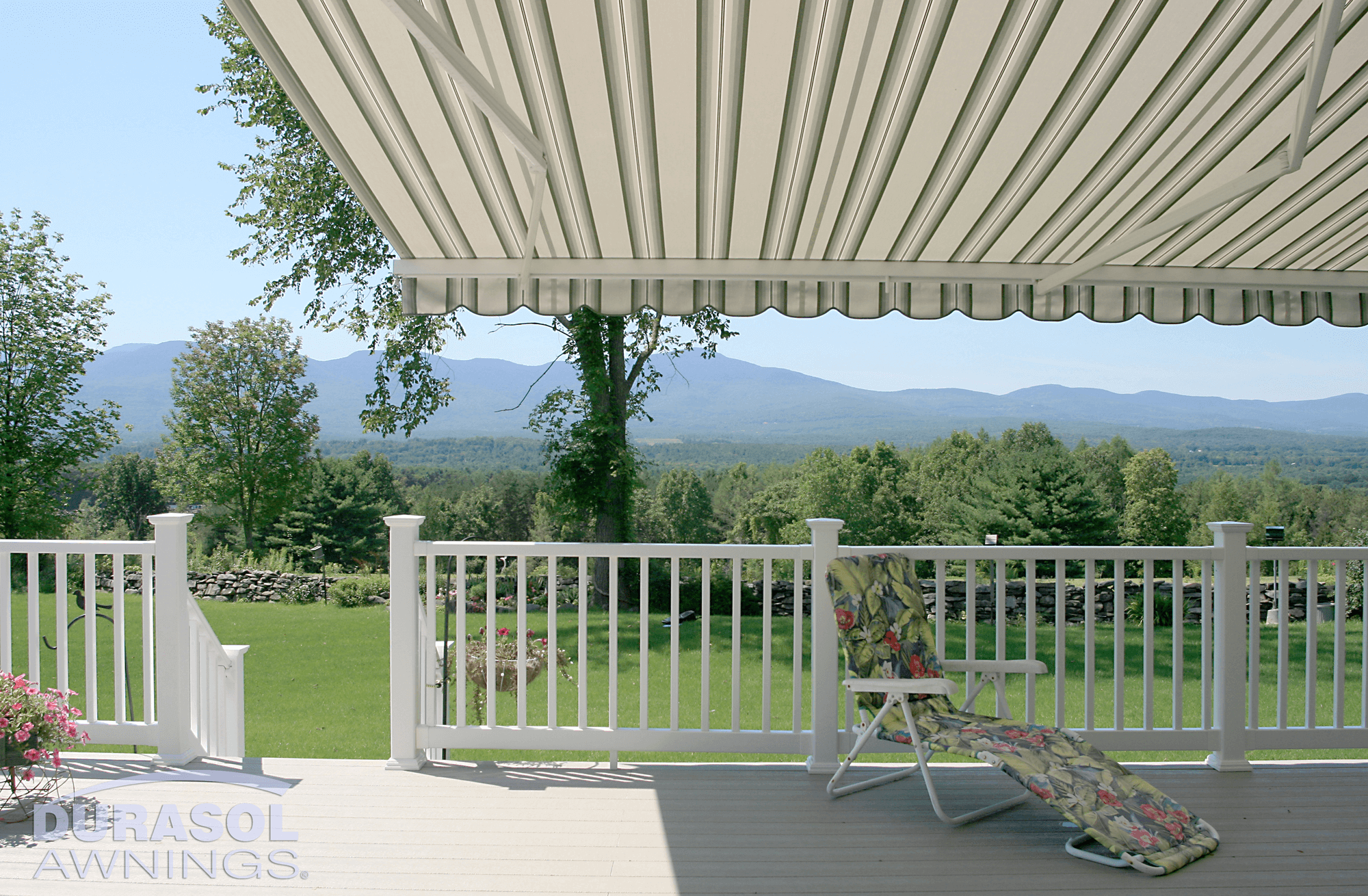 A porch deck with a white railing, striped awning, and a floral lounge chair overlooking a green yard, trees, and mountains in the background.