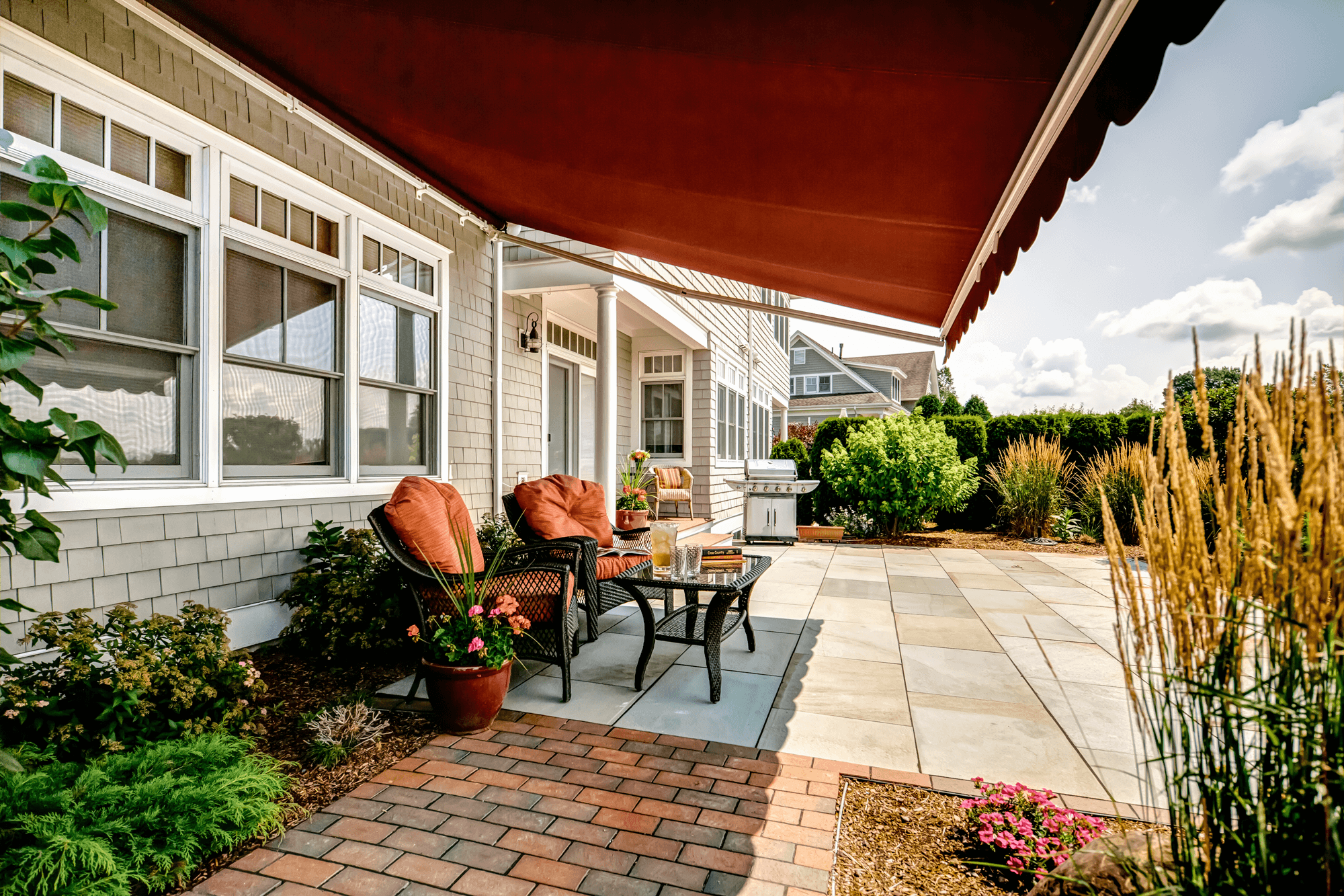 A backyard patio with outdoor chairs, a small table, a potted plant, a barbecue grill, lush green bushes, and decorative plants under a red patio umbrella on a sunny day.