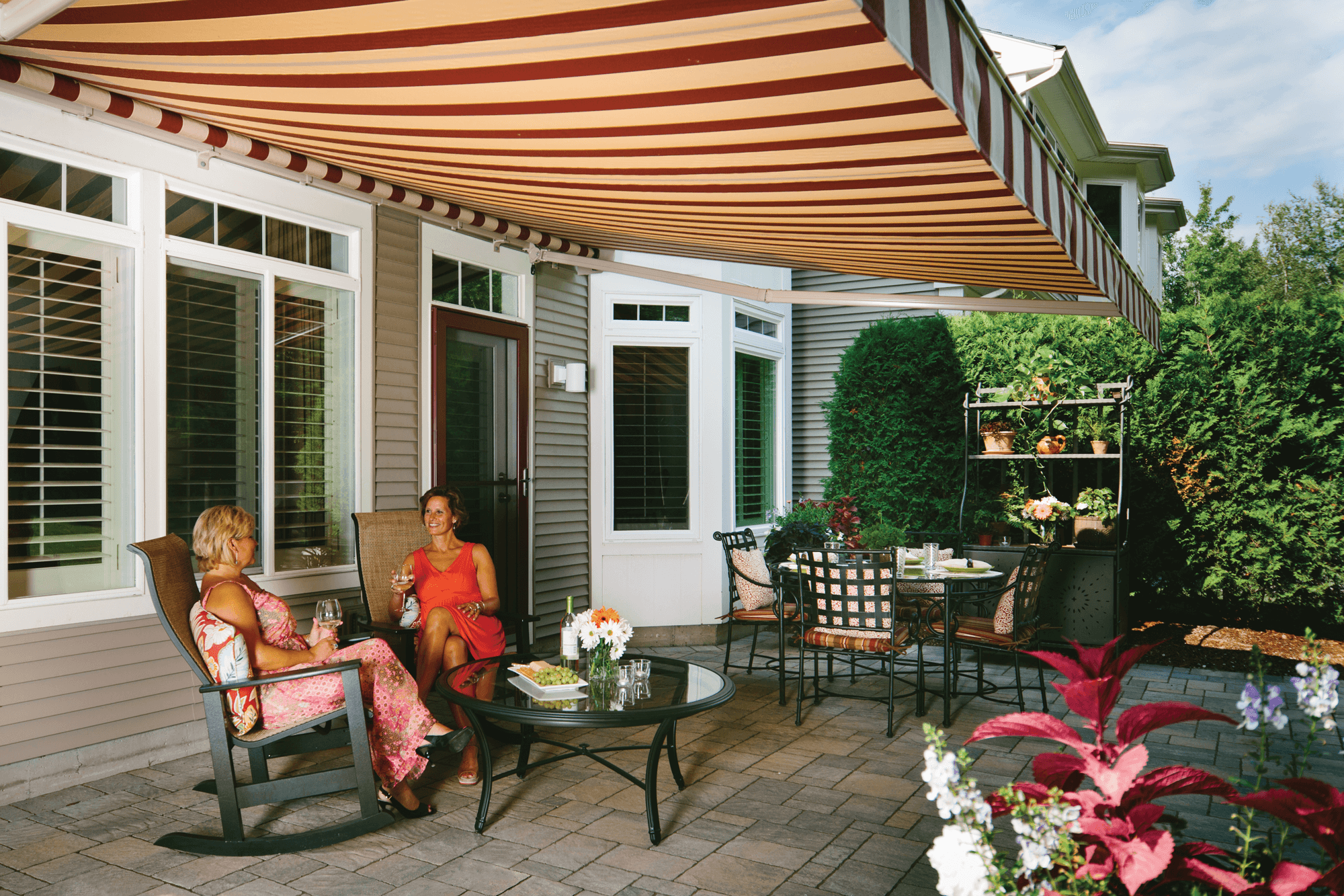 Two women sitting and chatting under a striped beige and red patio awning on a backyard patio, with potted plants and outdoor furniture around.