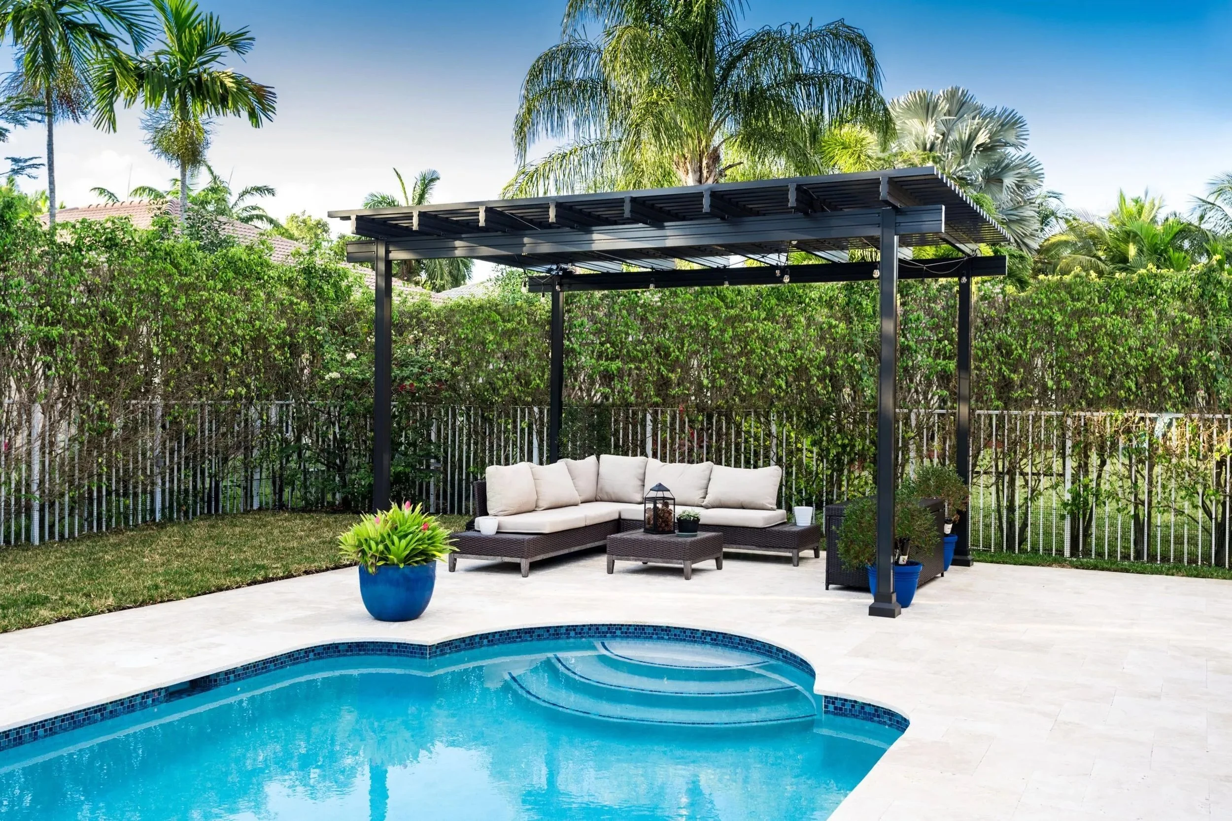 Backyard with swimming pool, outdoor sofa with cushions under a pergola, potted plants, and lush green trees.