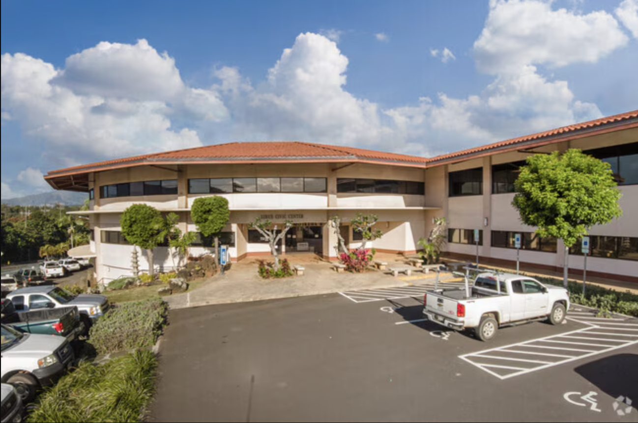Front view of a two-story medical clinic building with landscaped front, several trees, parked cars, and a clear sky with clouds.