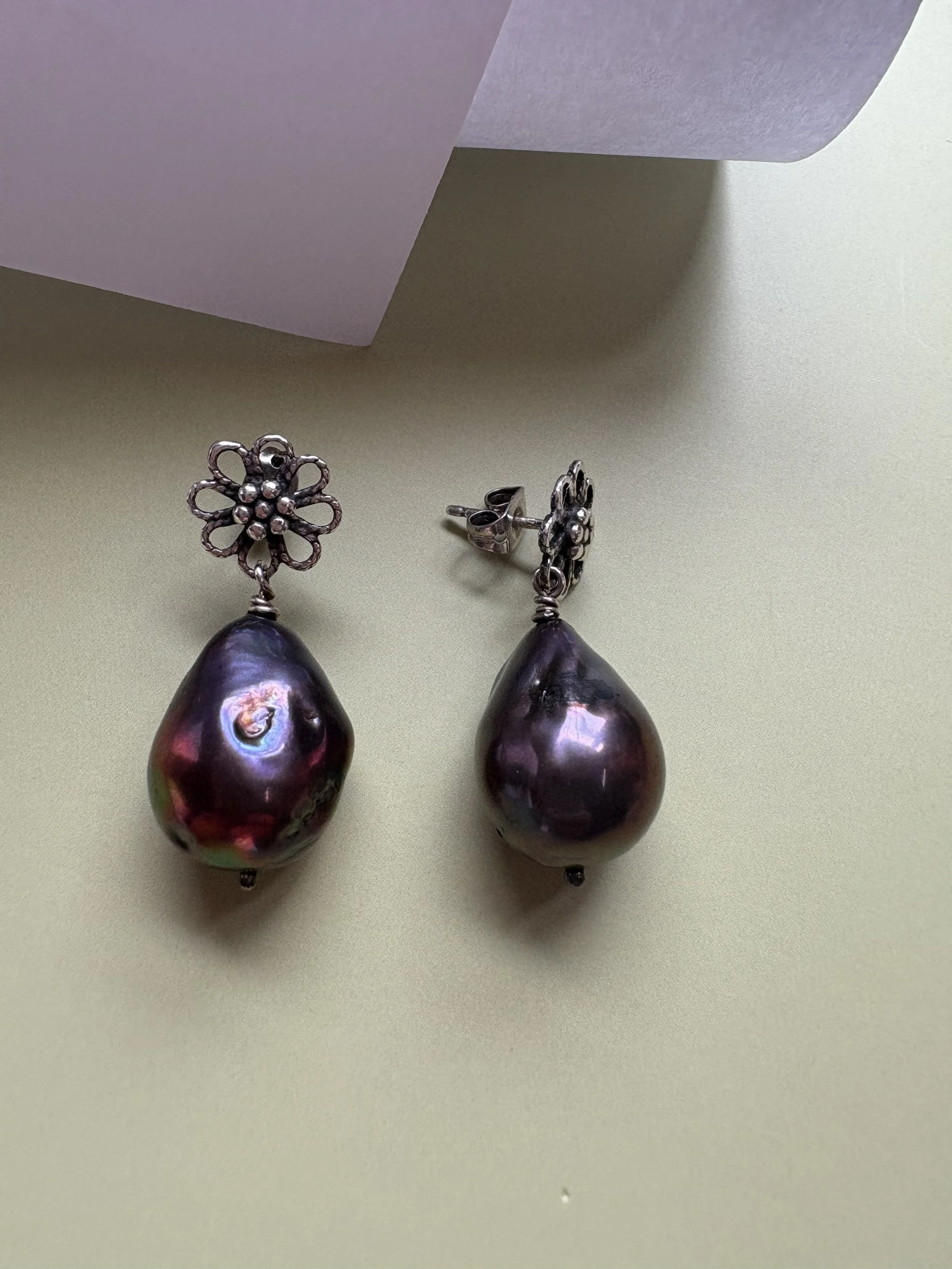 Pair of earrings with dark, irregularly shaped pearls and silver floral designs on a light green surface, with a white paper in the background.