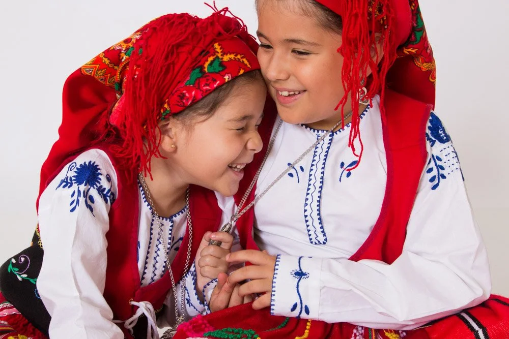 Two young girls in traditional Eastern European folk costumes smiling and laughing, close together, dressed in brightly colored embroidery clothing and red head coverings.