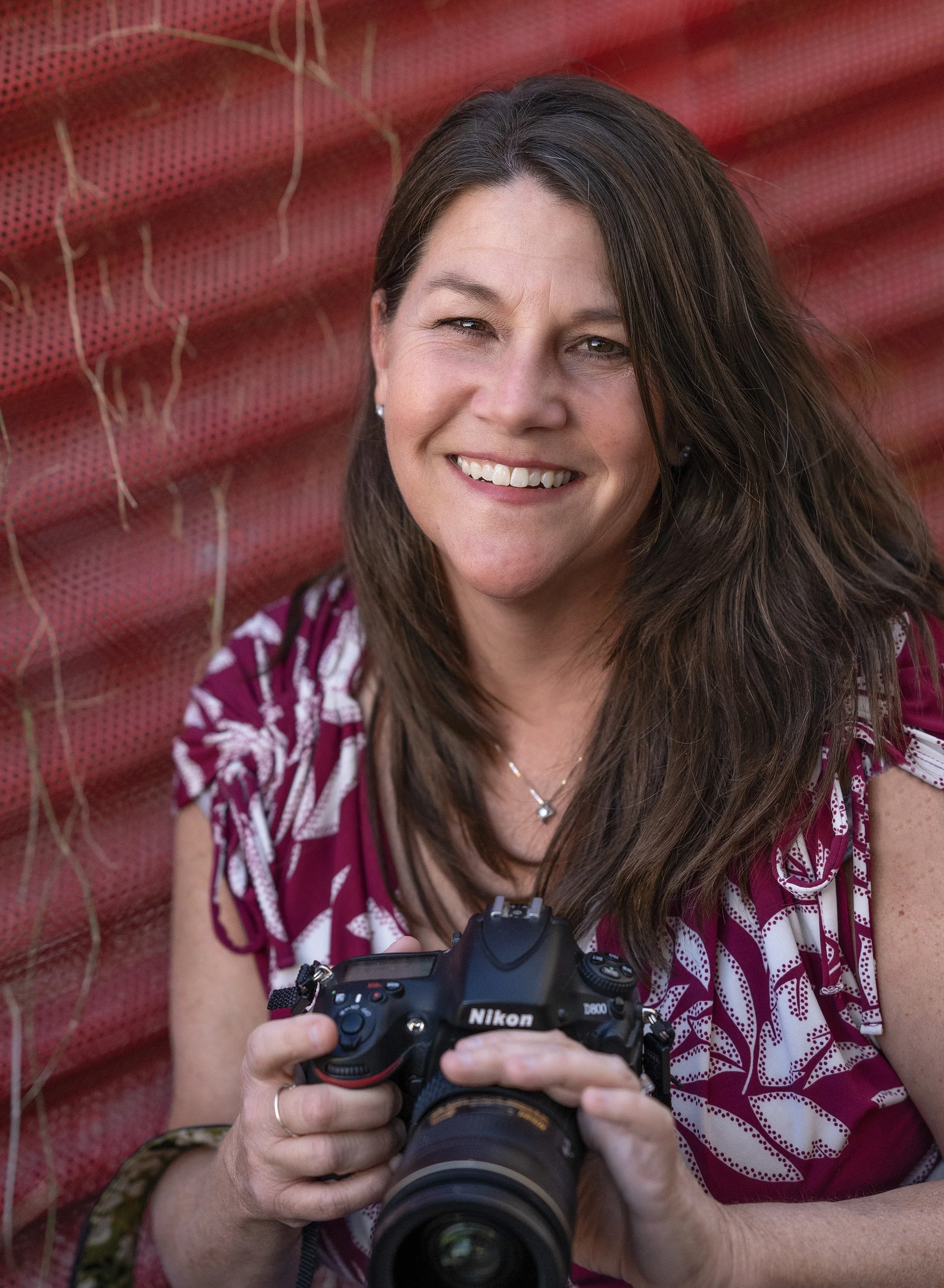 Photographer Kim Lloyd, holding a Nikon camera, smiling, and wearing a pink and white patterned shirt, in front of a red textured background.