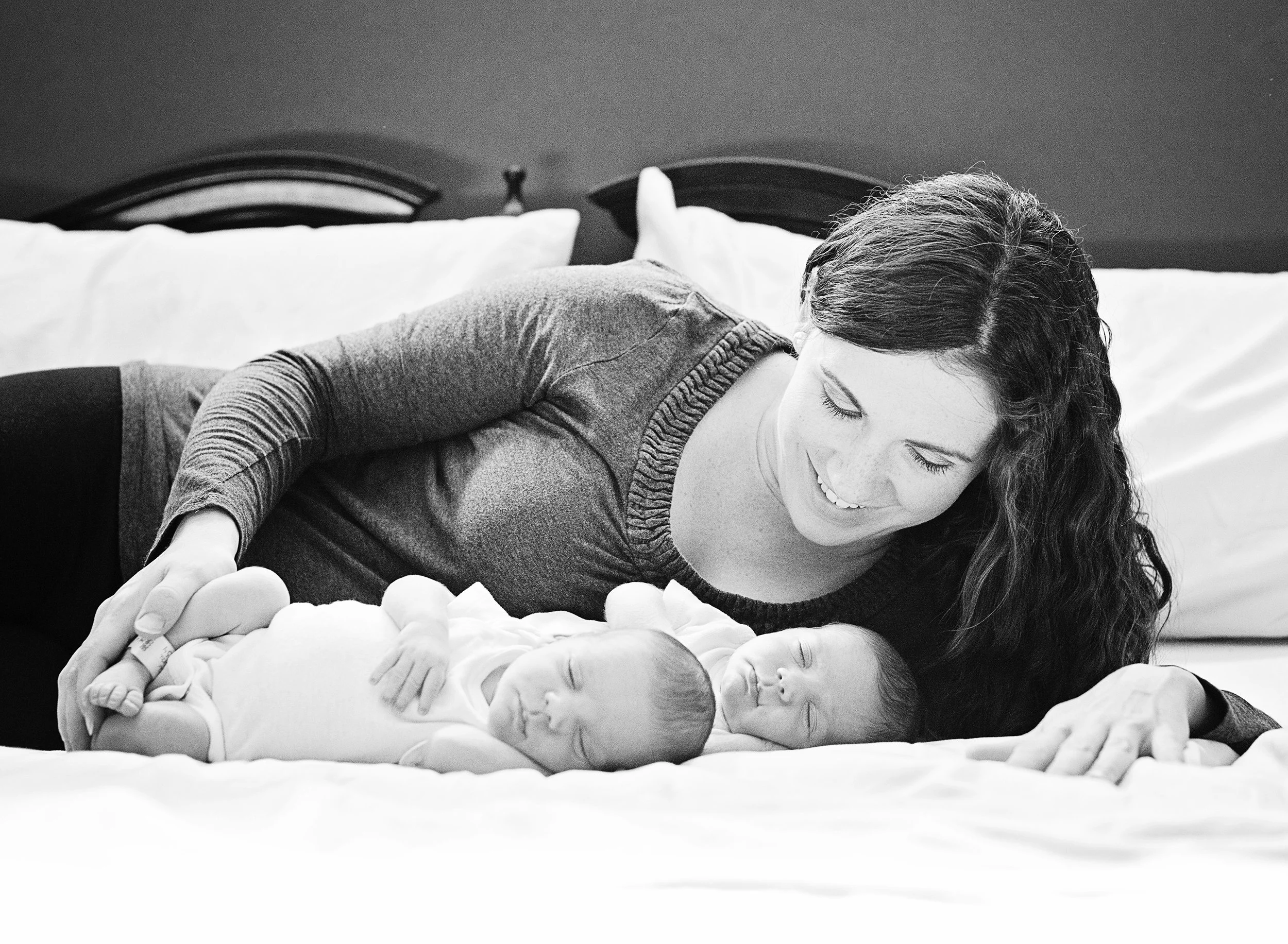 Black and white photo of a woman with long hair lying on a bed, smiling, with two newborn babies, one lying on her back and the other lying face up on the bed at a newborn session in Tampa.