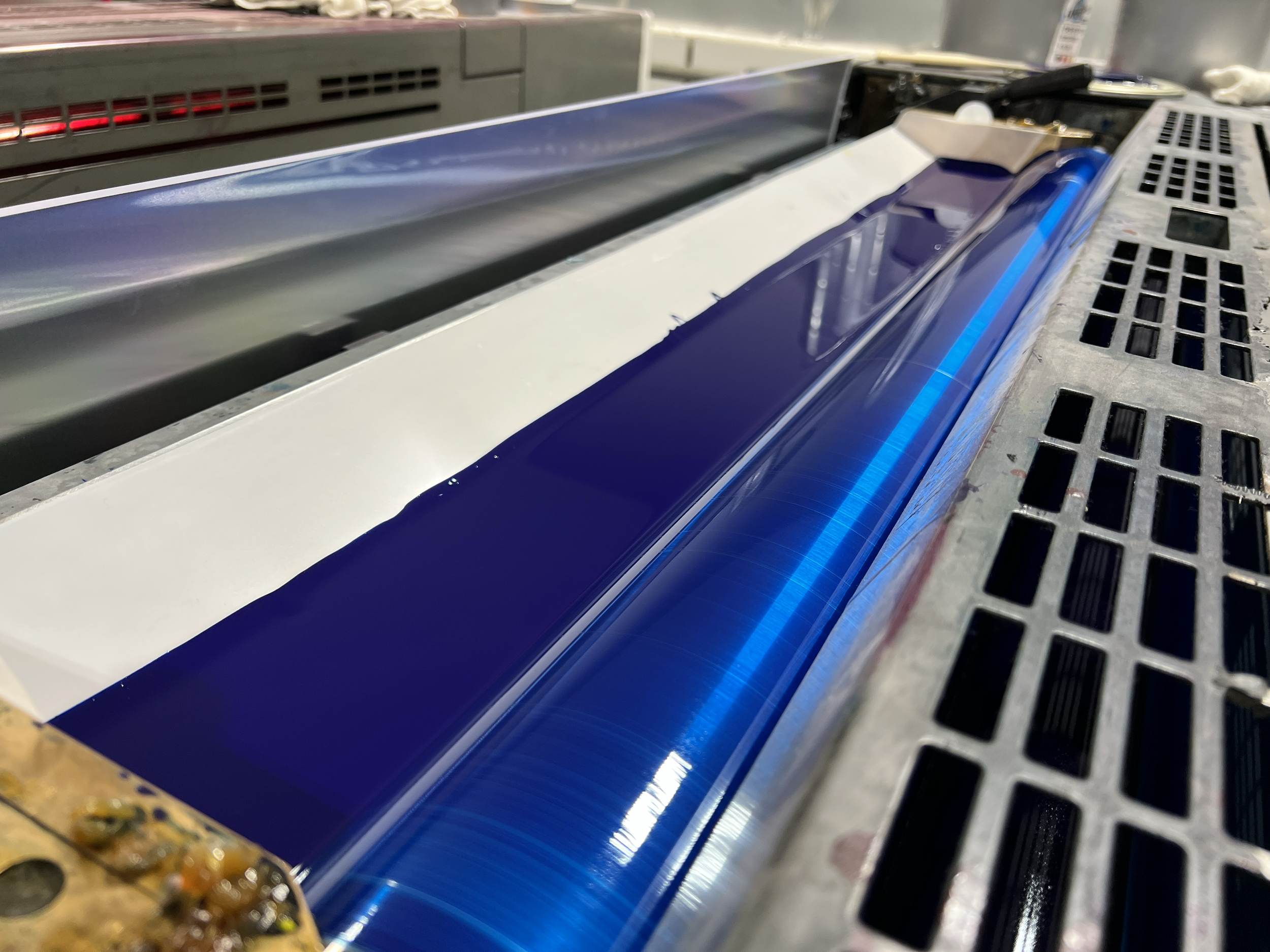 Close-up of a printing or manufacturing process with a shiny, blue, metallic surface being processed on a machine with ventilation grates.