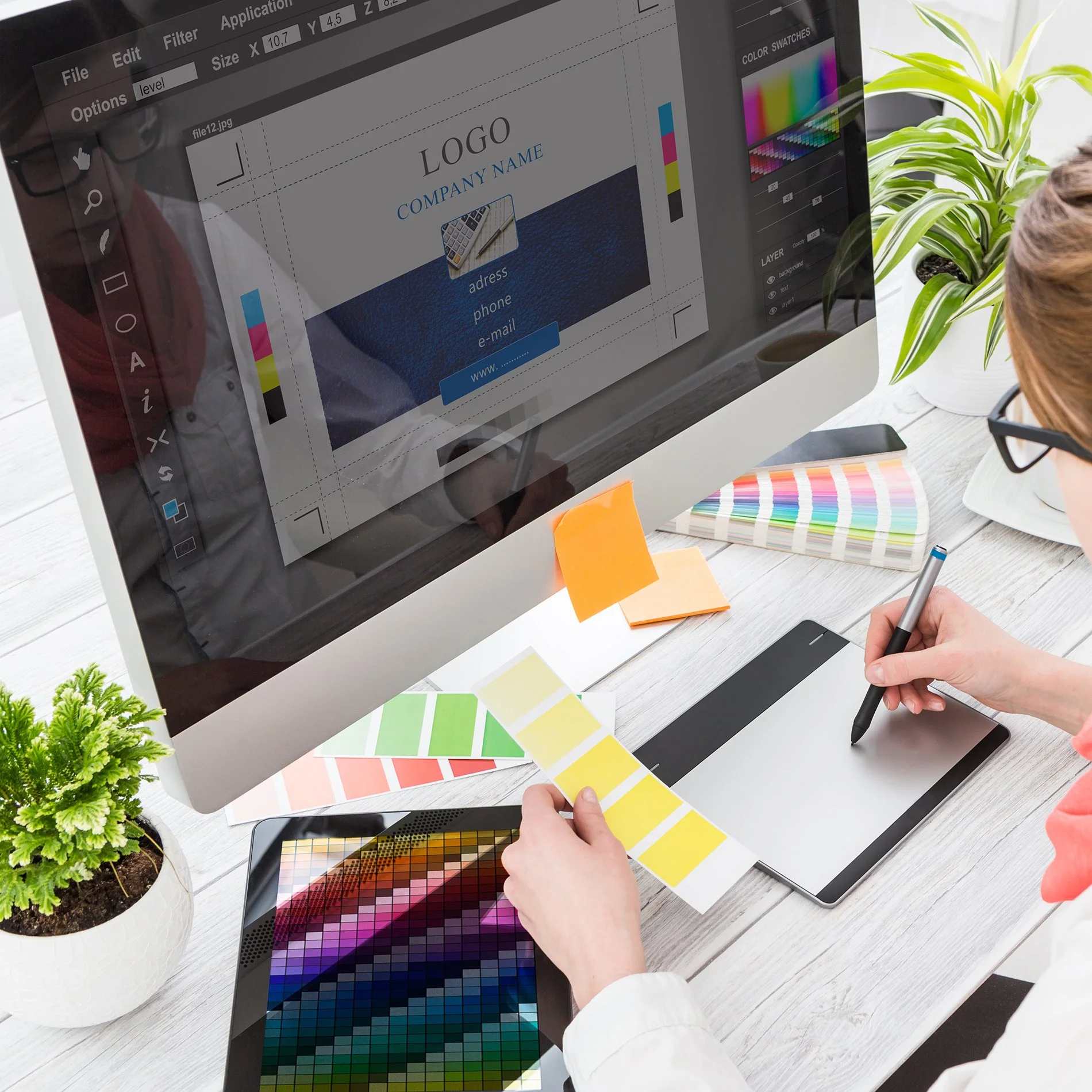 A person working on graphic design for a website on a desktop computer, with color swatches and design tools on a white wooden desk, and potted plants nearby.