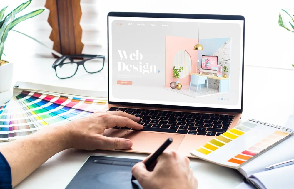Person designing a website on a laptop, surrounded by color swatches and a sketchbook, with desk accessories like glasses and plants in the background.