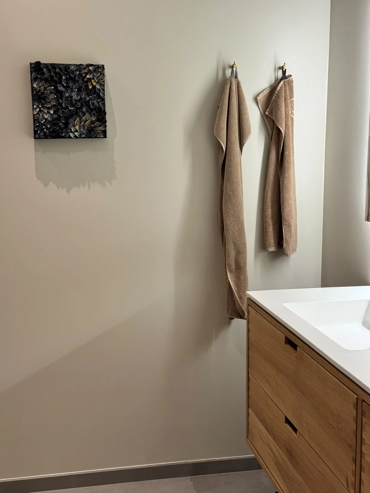 Two beige towels hanging on hooks on a white wall in a bathroom, with part of a wooden cabinet and white countertop visible in the lower right corner, and a black decorative art piece on the wall to the left.