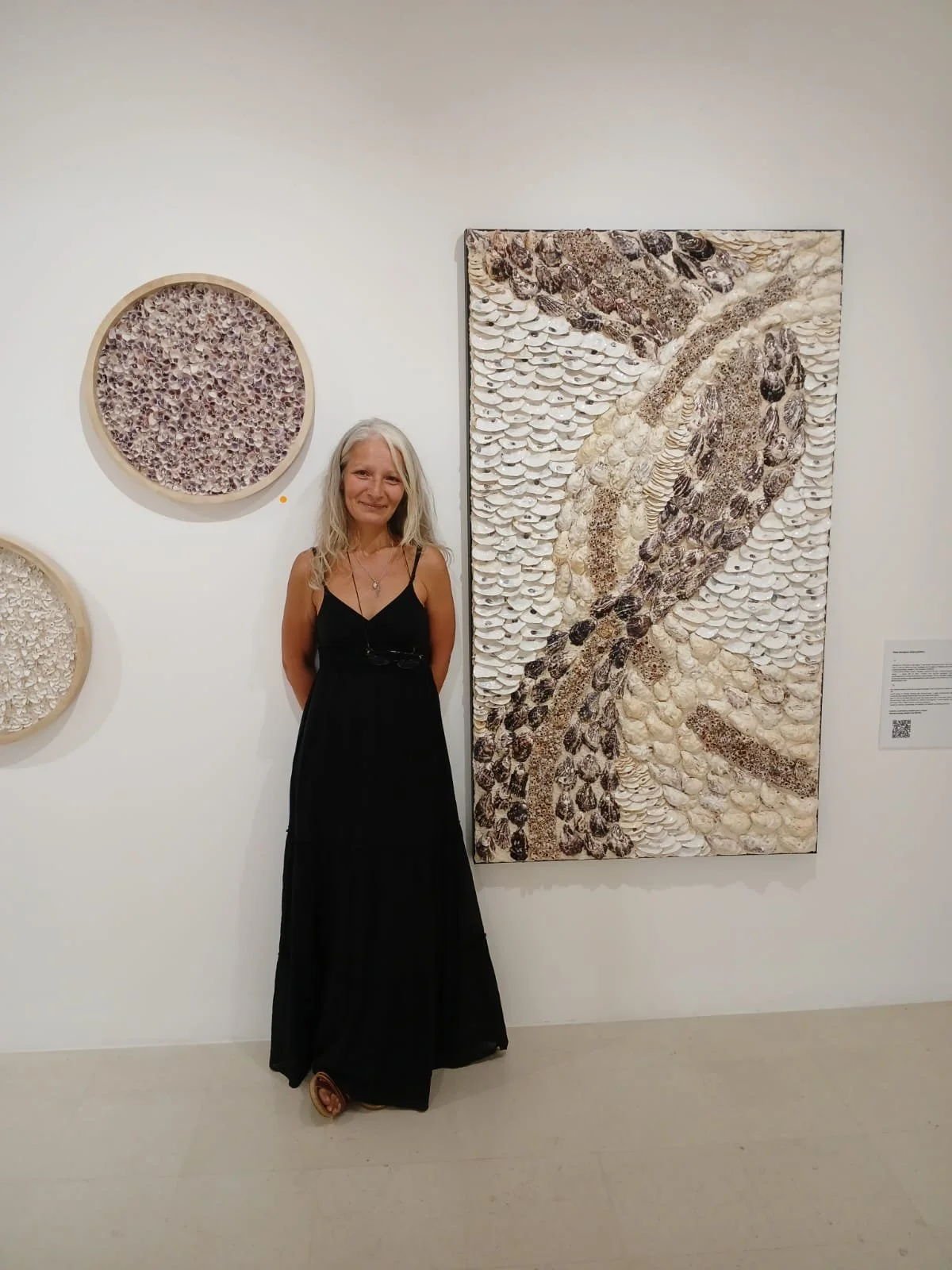woman artist dressed in a black dress, with long grey hair, standing by wall exhibition, next to a large and a round shell artworks. 