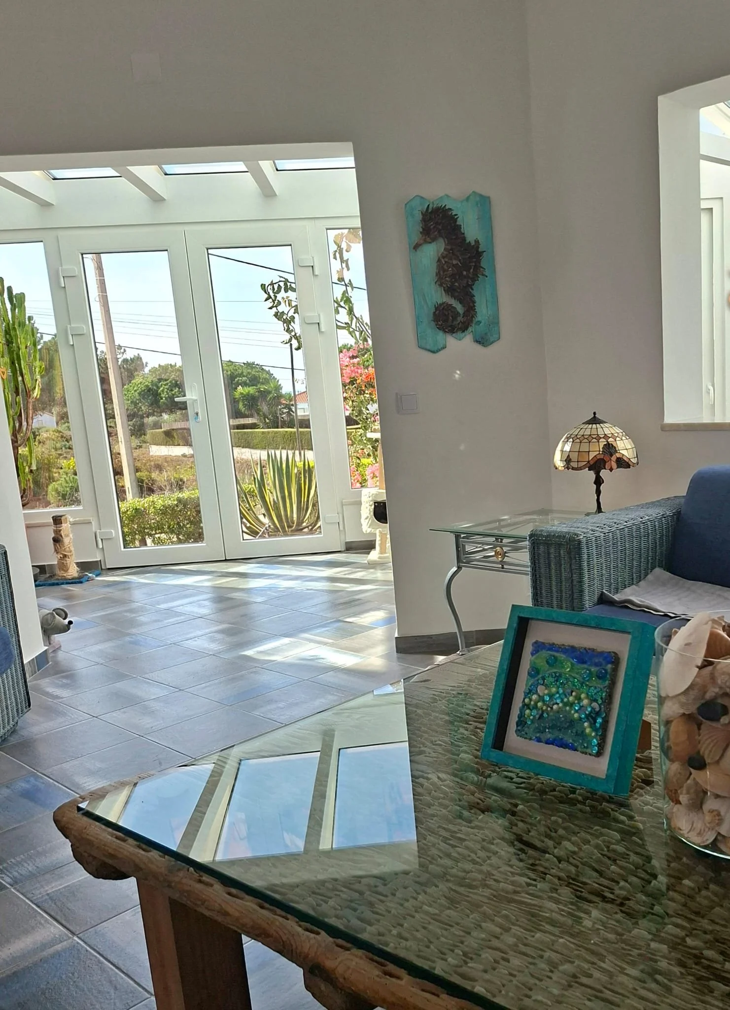 A bright living room with large glass doors leading outside, a tiled floor, colorful decorations, a lamp, and a wicker chair.