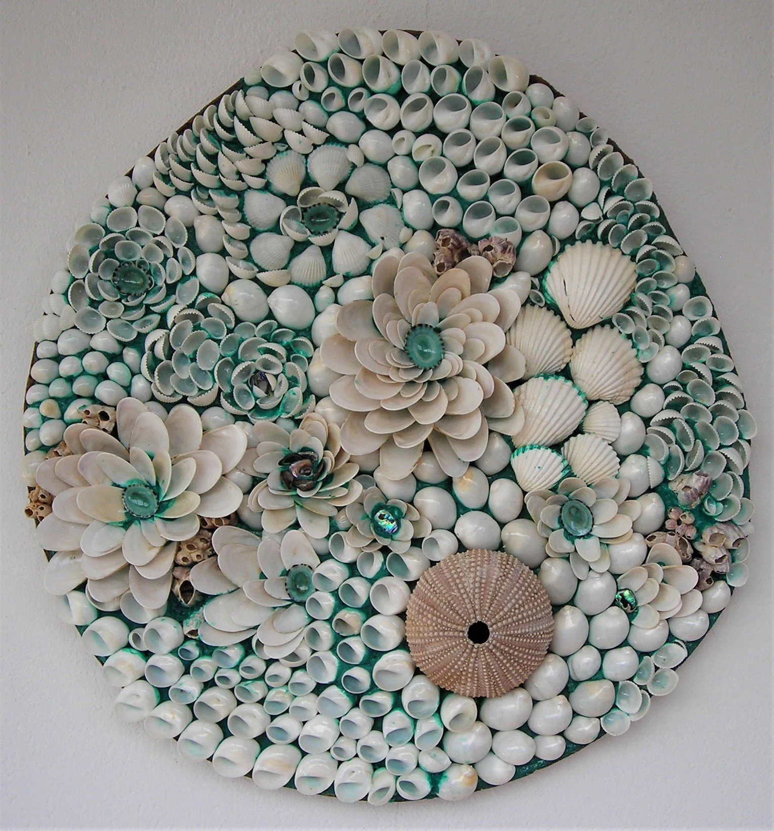 A round mirror with a wooden frame, and a decorative wall art piece made of seashells arranged in a floral pattern, on a white wall.