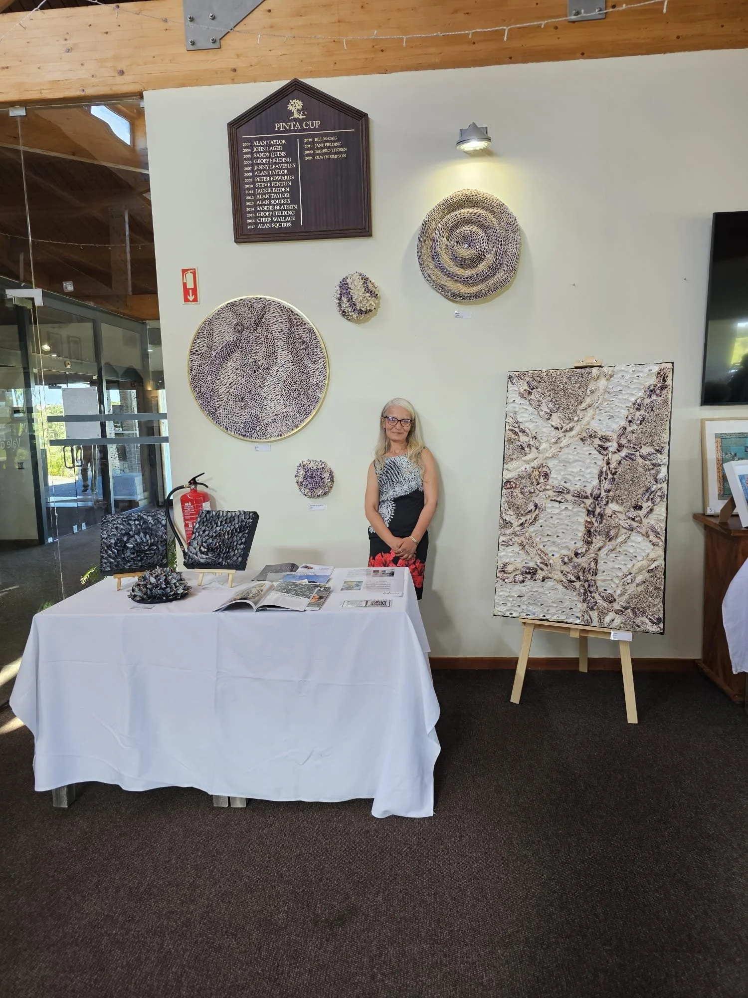 An indoor art exhibit with a table displaying art pieces made from shells, a woman standing behind the table, and various shell art on the wall behind her, including framed shell designs and woven shell sculptures.