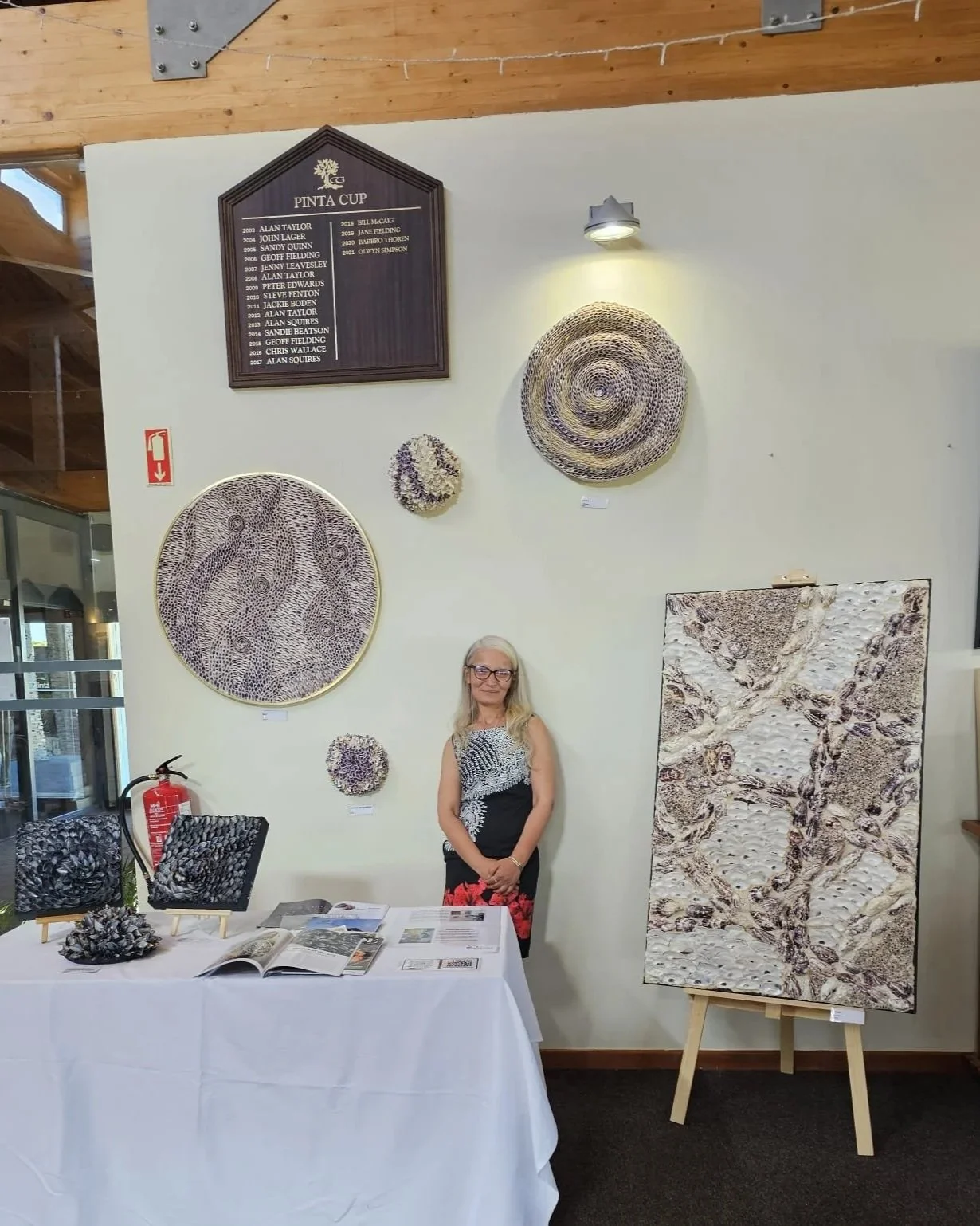 An indoor art exhibit with a table displaying art pieces made from shells, a woman standing behind the table, and various shell art on the wall behind her, including framed shell designs and woven shell sculptures.