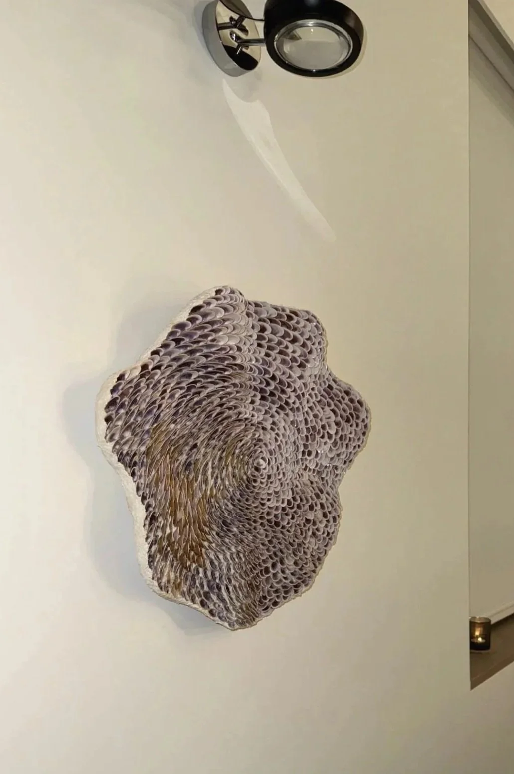 Decorative wavey-shaped wall art made of rolled paper on a white wall, with a black wall-mounted light fixture above it.