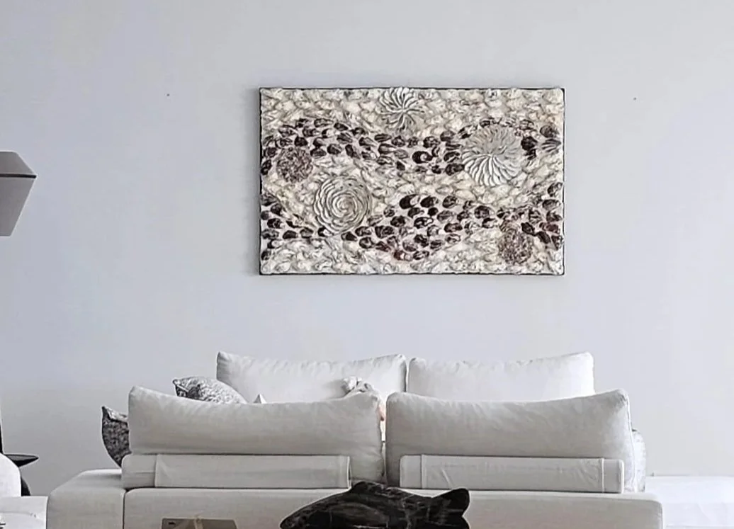 A living room with a light-colored sofa, several white pillows, and a decorative abstract artwork made of oyster shells with swirling patterns and swirls hanging above the sofa.