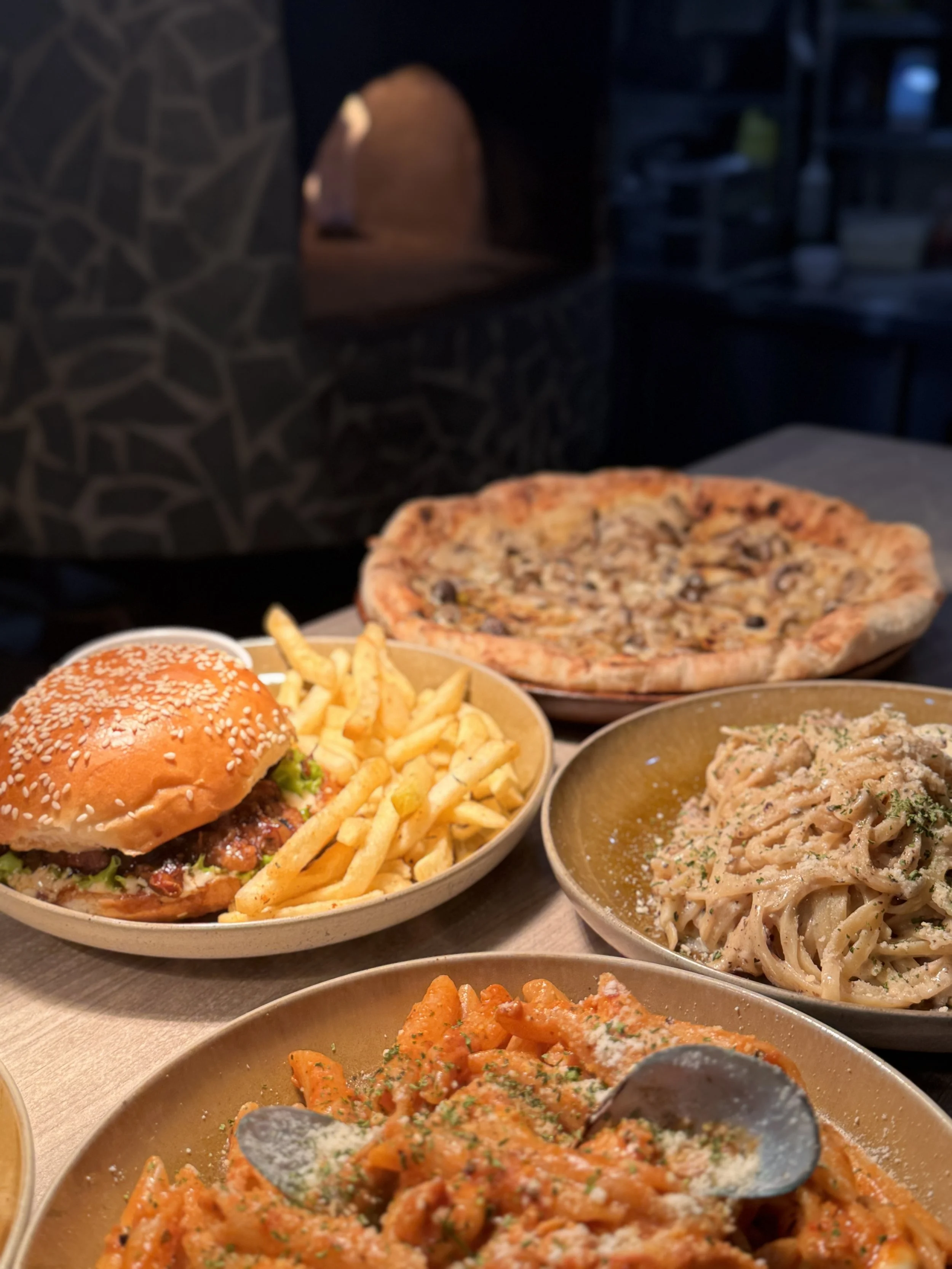 A variety of Italian and American dishes on a table including a pizza, a hamburger with fries, and plates of pasta, with a wood-fired oven in the background.