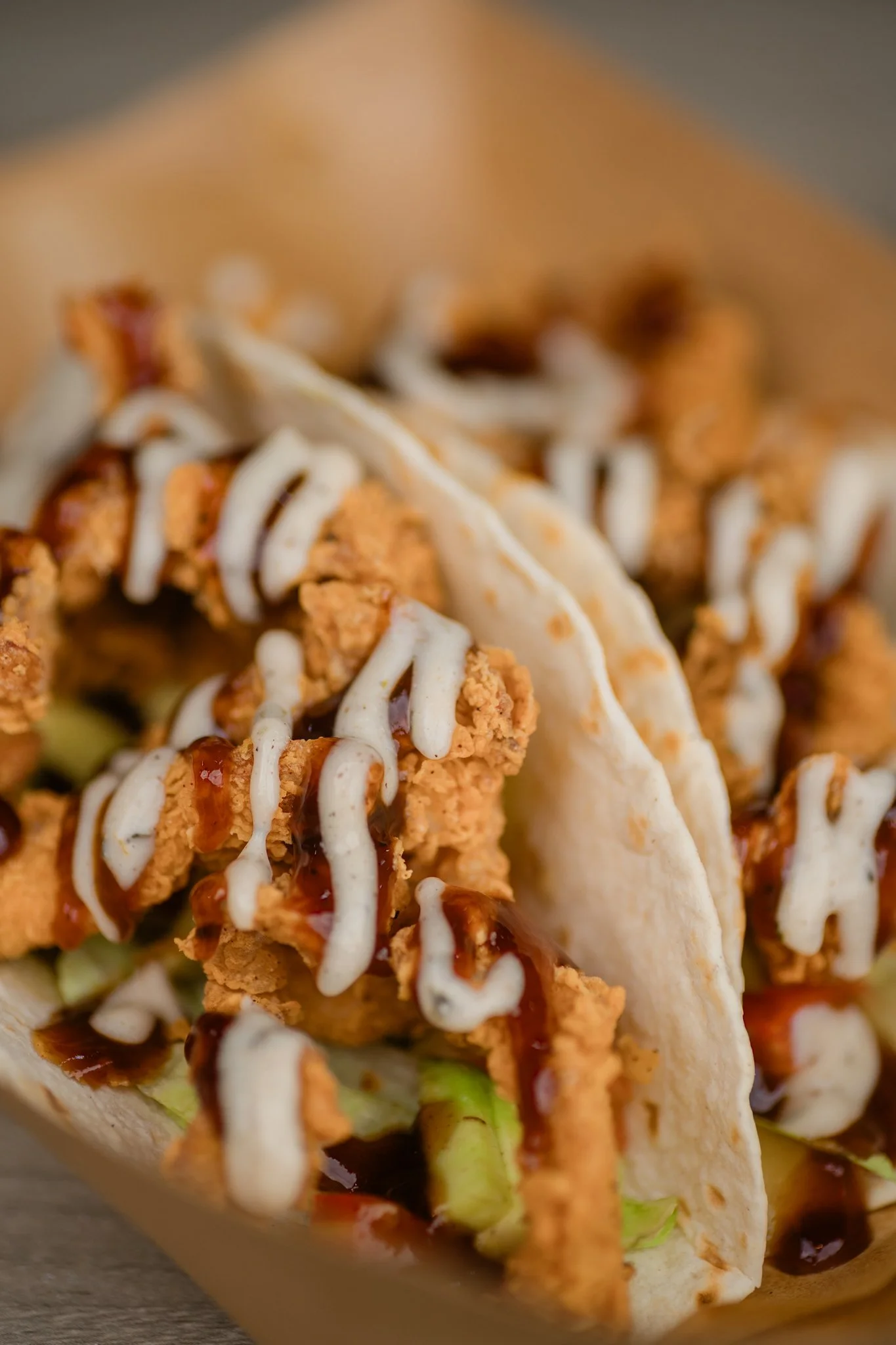 Close-up of two tacos filled with fried chicken, lettuce, drizzled with white sauce and dark sauce, in soft flour tortillas.