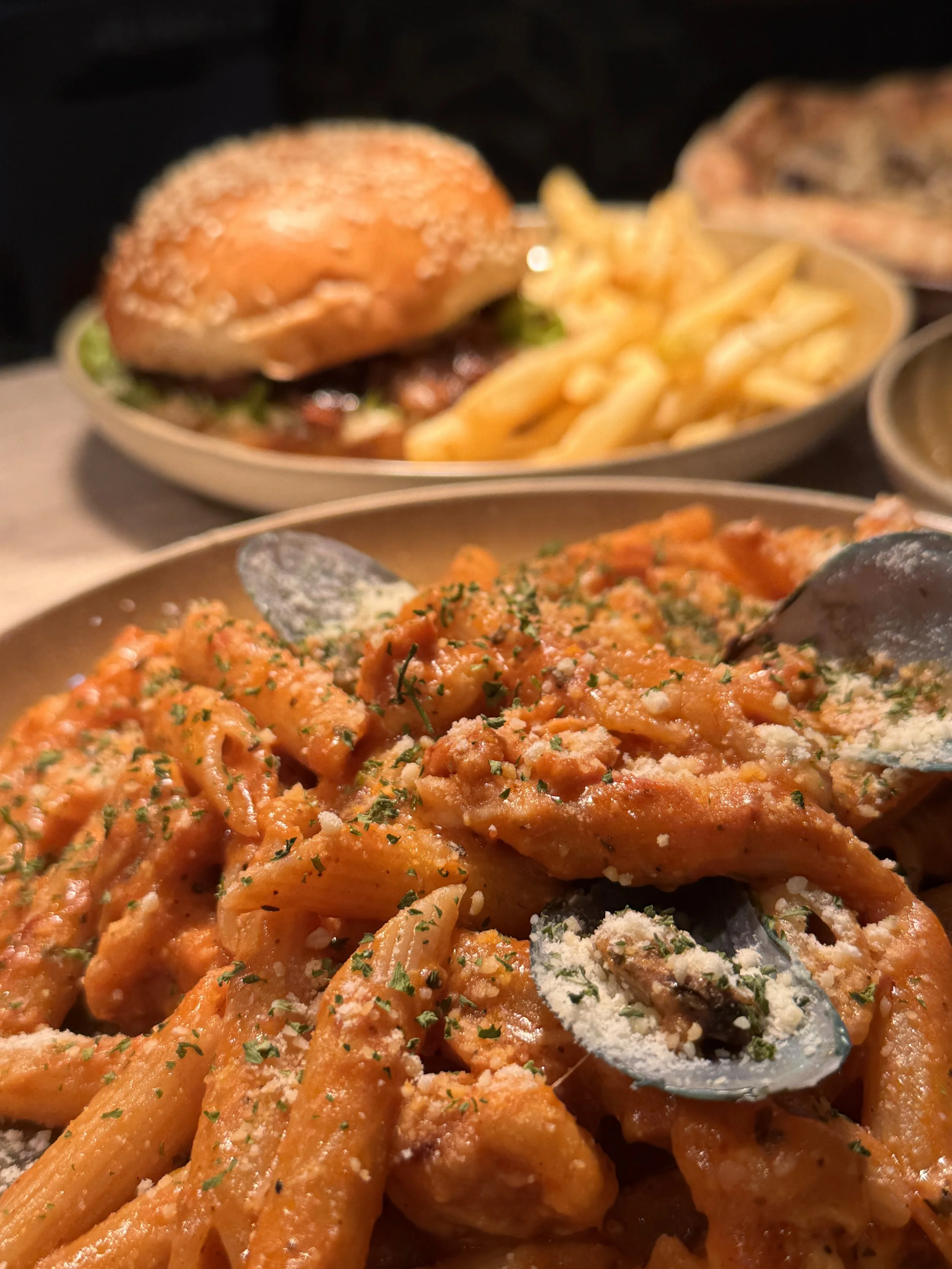 Close-up of a plate of pasta with tomato sauce, garnished with grated cheese and herbs. In the background, a burger with fries and another dish are visible.