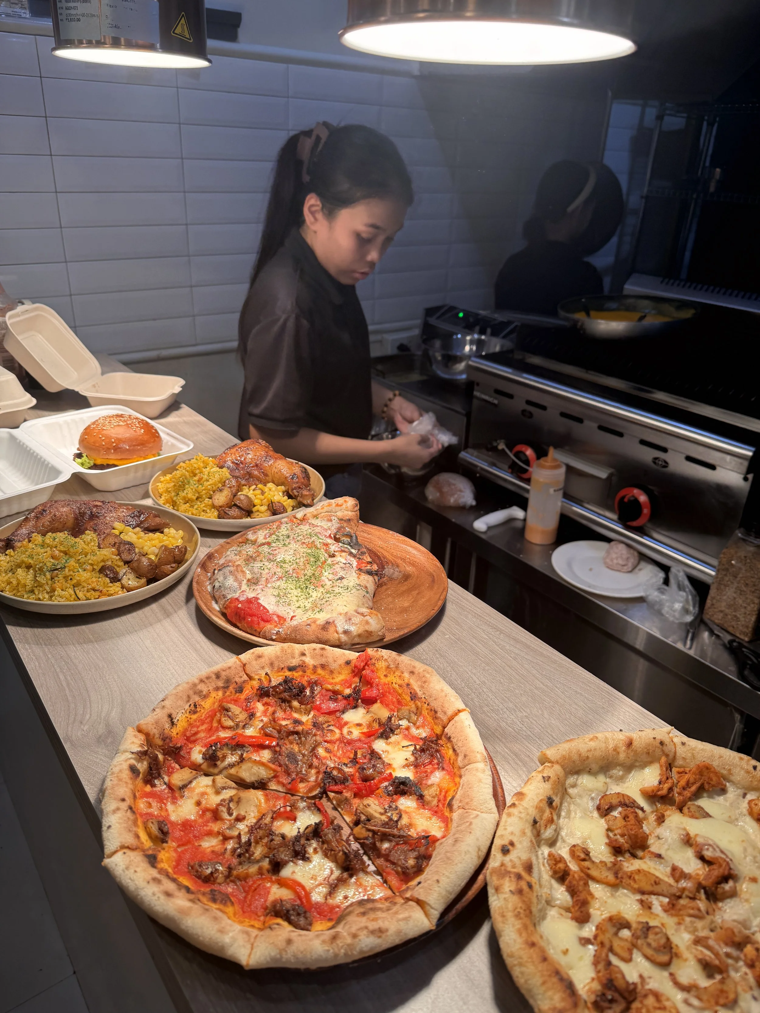 A woman preparing food in a kitchen surrounded by multiple pizzas and dishes on a counter.