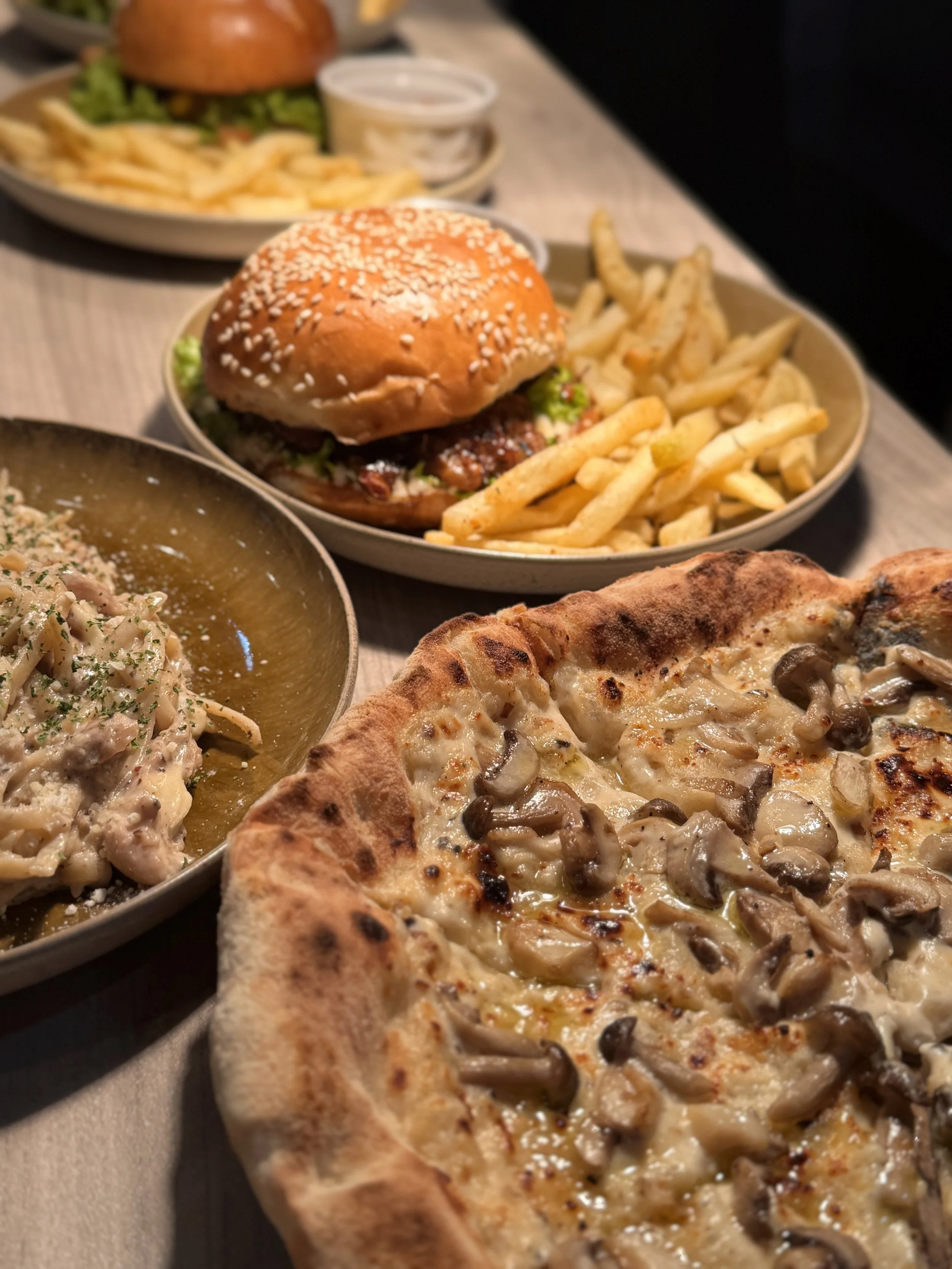 Close-up of a restaurant meal featuring a mushroom and cheese pizza, a pasta dish, a cheeseburger with fries, and a side of salad and dressing on a wooden table.