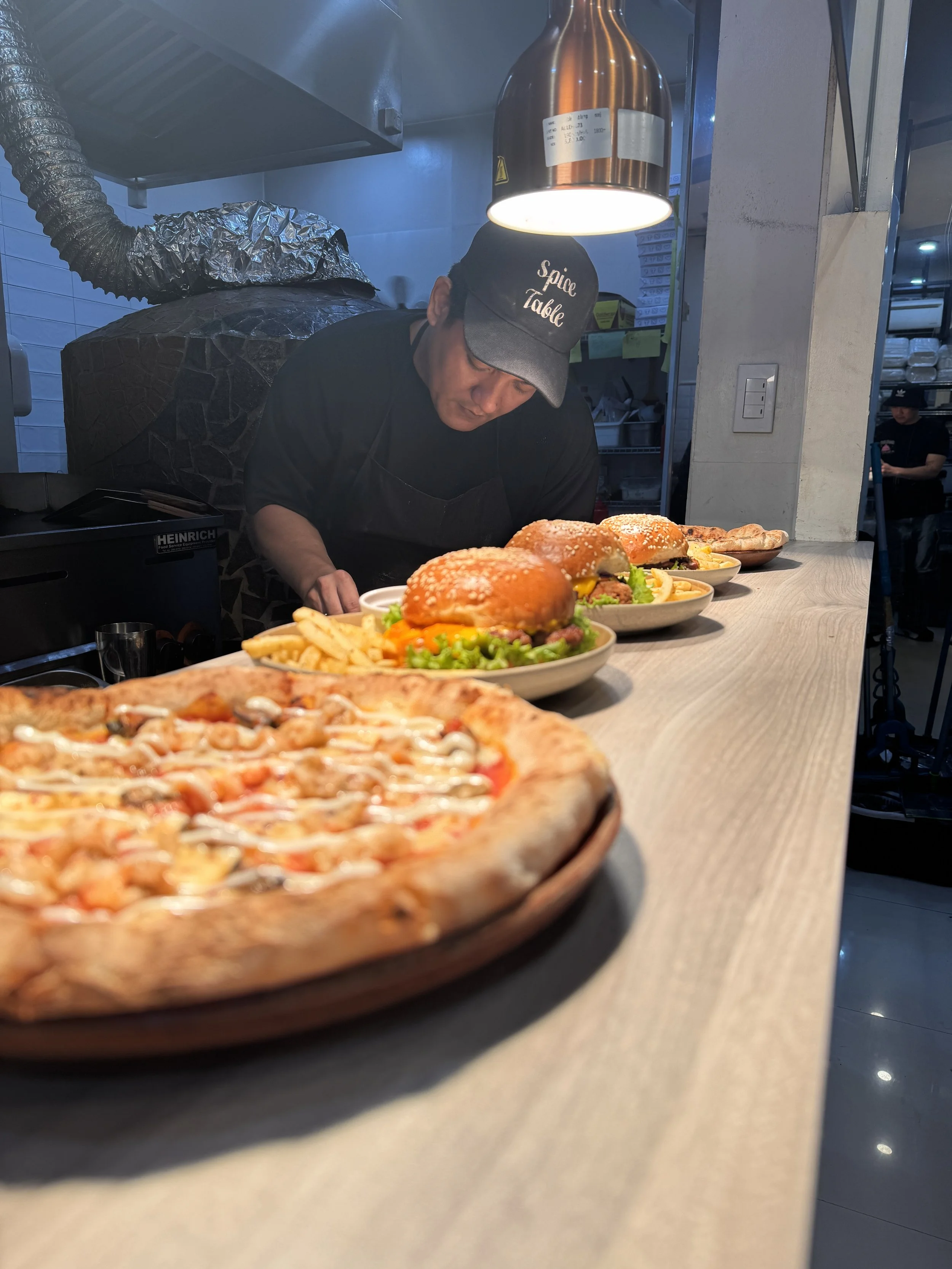 Pizza, burgers with fries, and a chef preparing food at a restaurant counter with a pizza in the foreground.