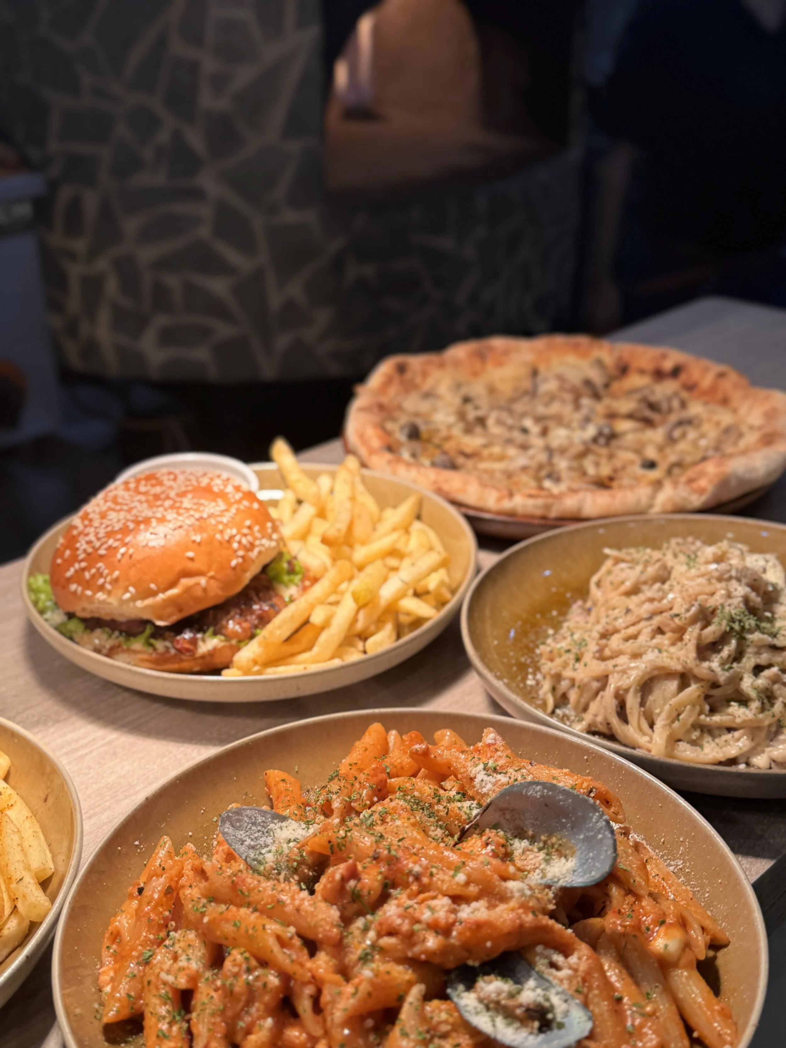 Assorted Italian and American dishes on a table, including a pasta dish, a pizza, a burger with fries, and a plate of pasta with sauce.