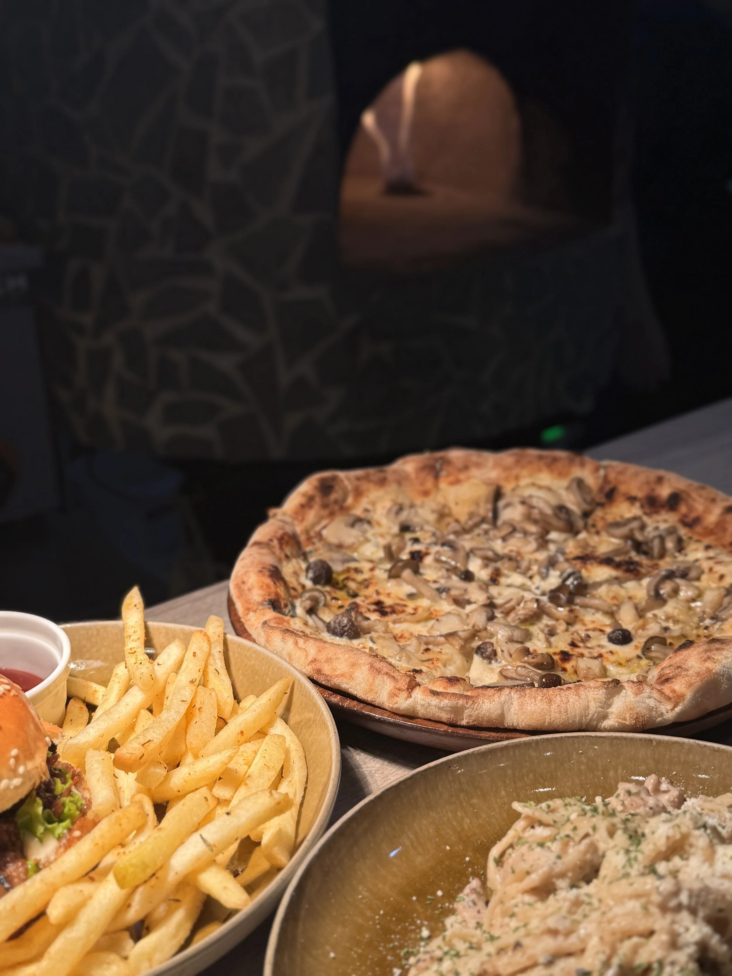 A pizza topped with mushrooms and black olives, a bowl of French fries with a side of ketchup and a small container of dipping sauce, and a bowl of creamy pasta with herbs on a table.