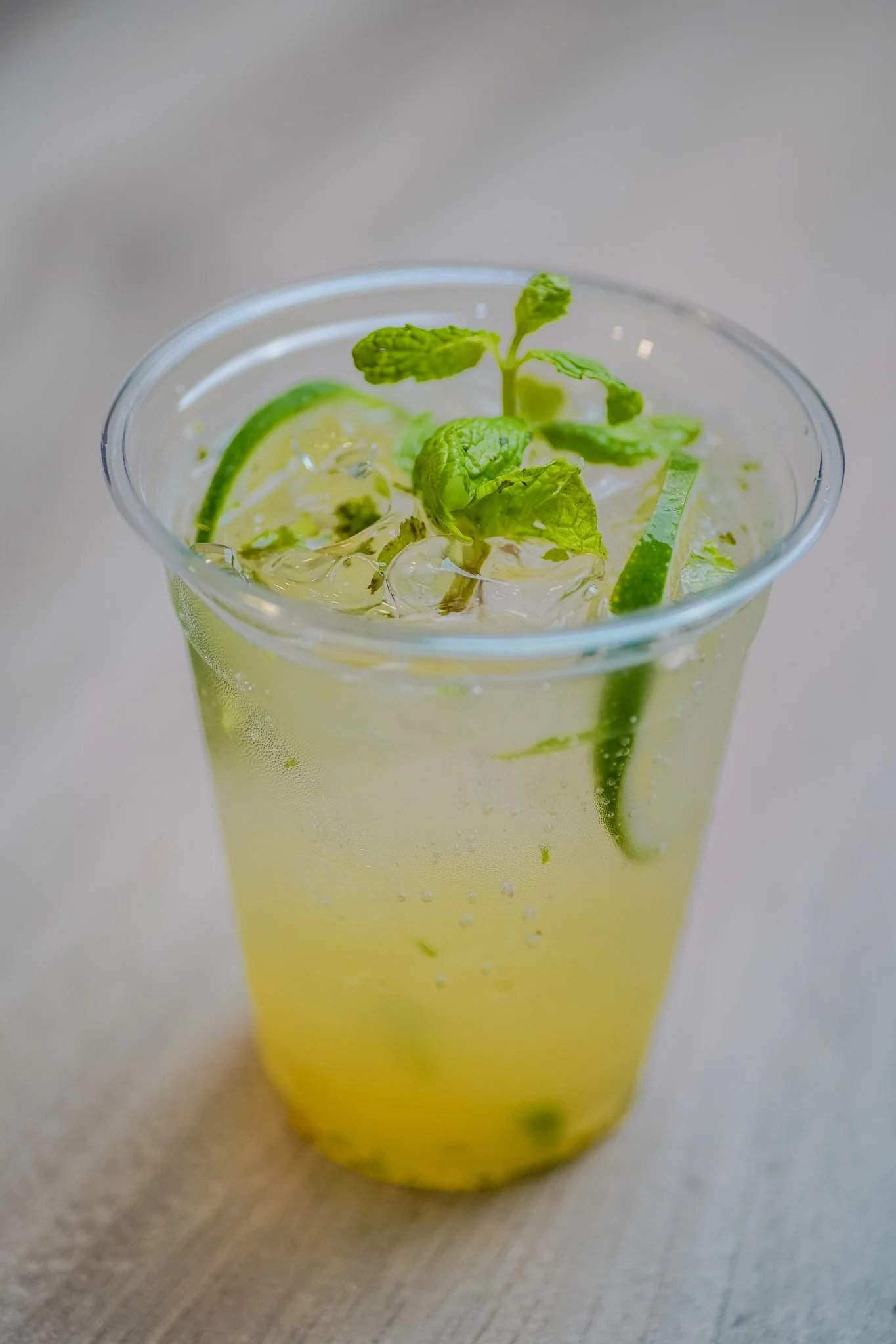 A clear plastic cup filled with a yellow-green beverage, ice cubes, cucumber slices, and fresh mint leaves, on a light-colored surface.