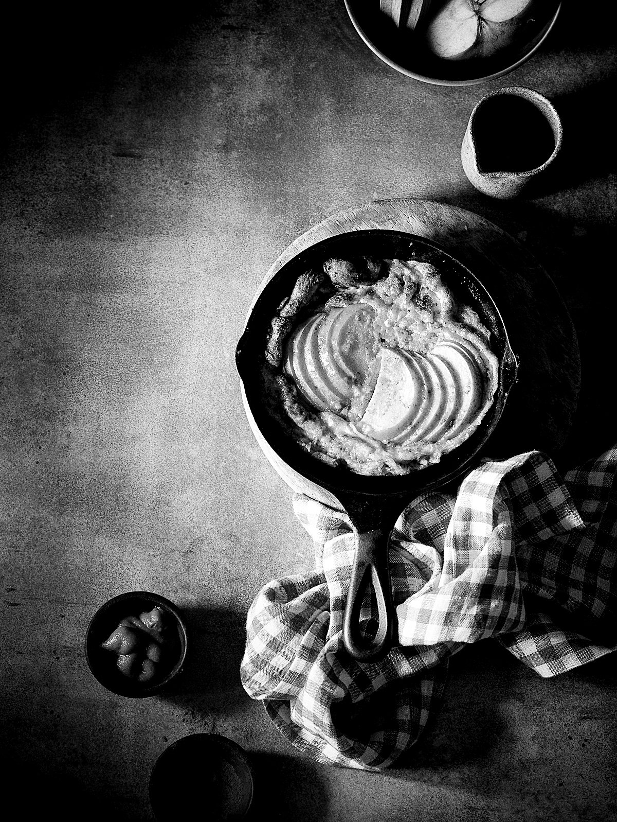 A cast iron skillet with scalloped potatoes and melted cheese, placed on a checkered cloth, on a textured surface. Surrounding bowls contain additional food items and a small container.