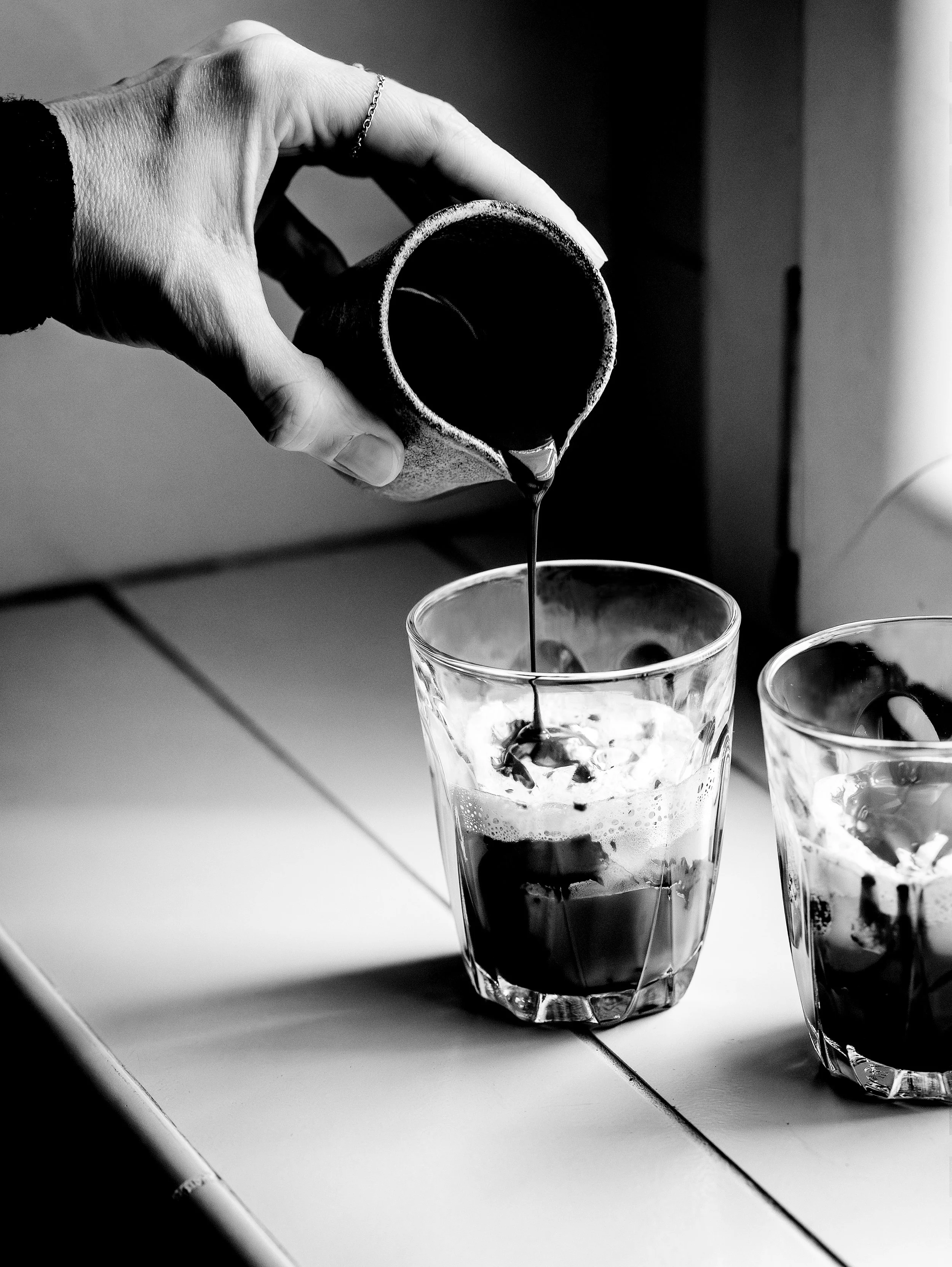 A black and white photo of a person's hand pouring coffee from a small pitcher into a glass filled with ice on a white tiled surface near a window.