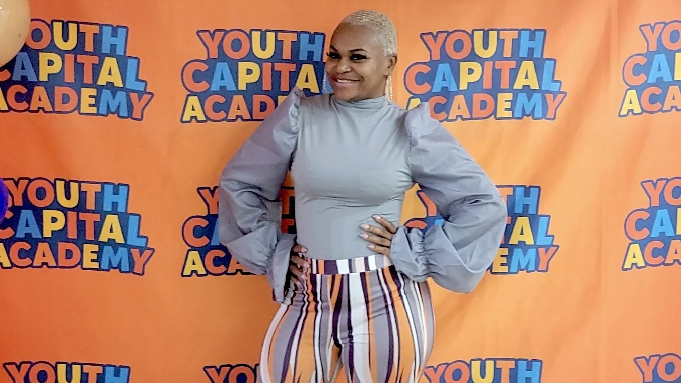 A woman with short blonde hair, smiling, wearing a gray puff-sleeve top and striped pants, standing in front of an orange backdrop with the logo "YOUTH CAPITAL ACADEMY" repeating in bright colors.