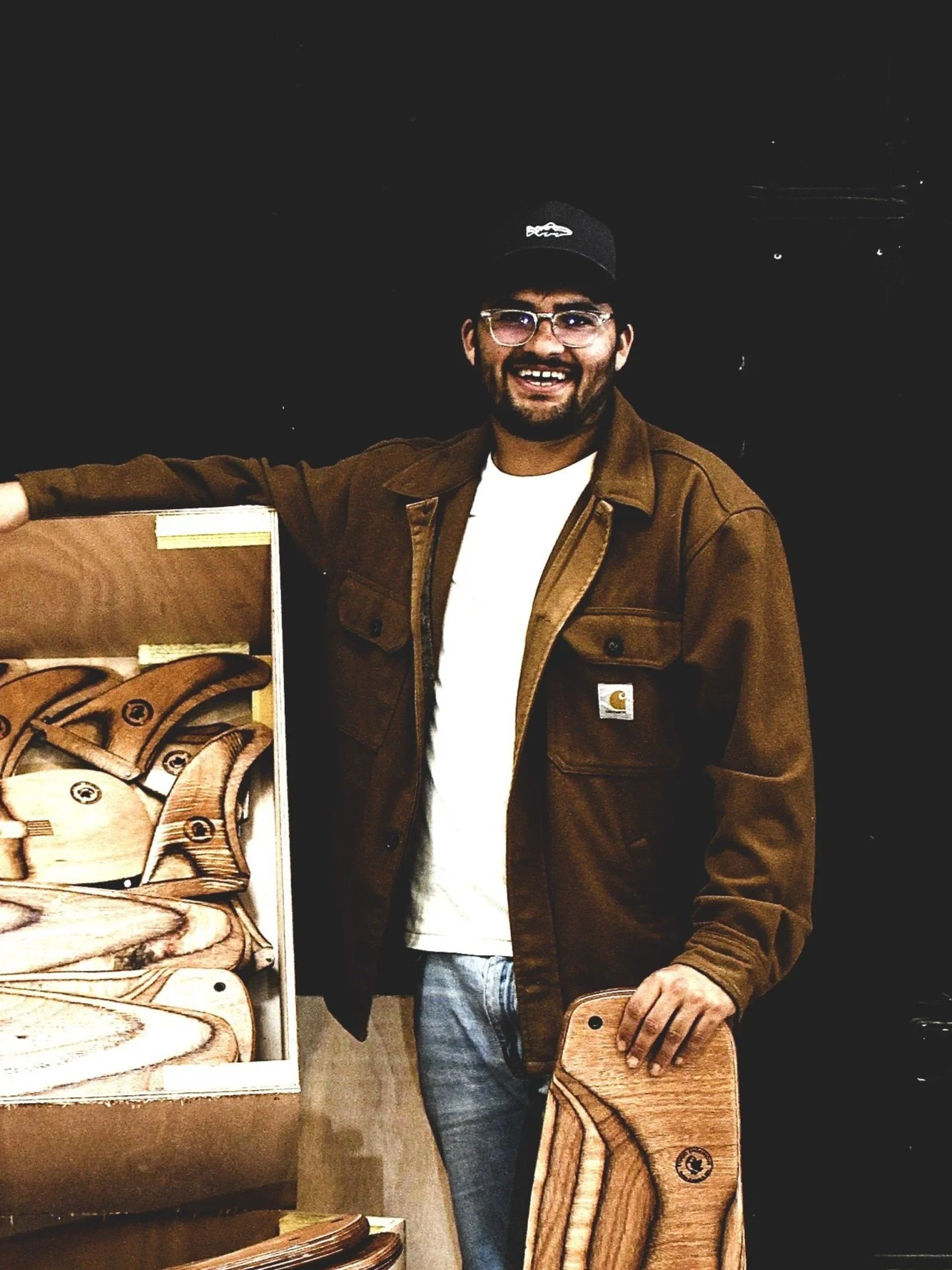 A man with glasses, a beard, and a baseball cap smiling while standing next to a display of wooden cutting boards and utensils.