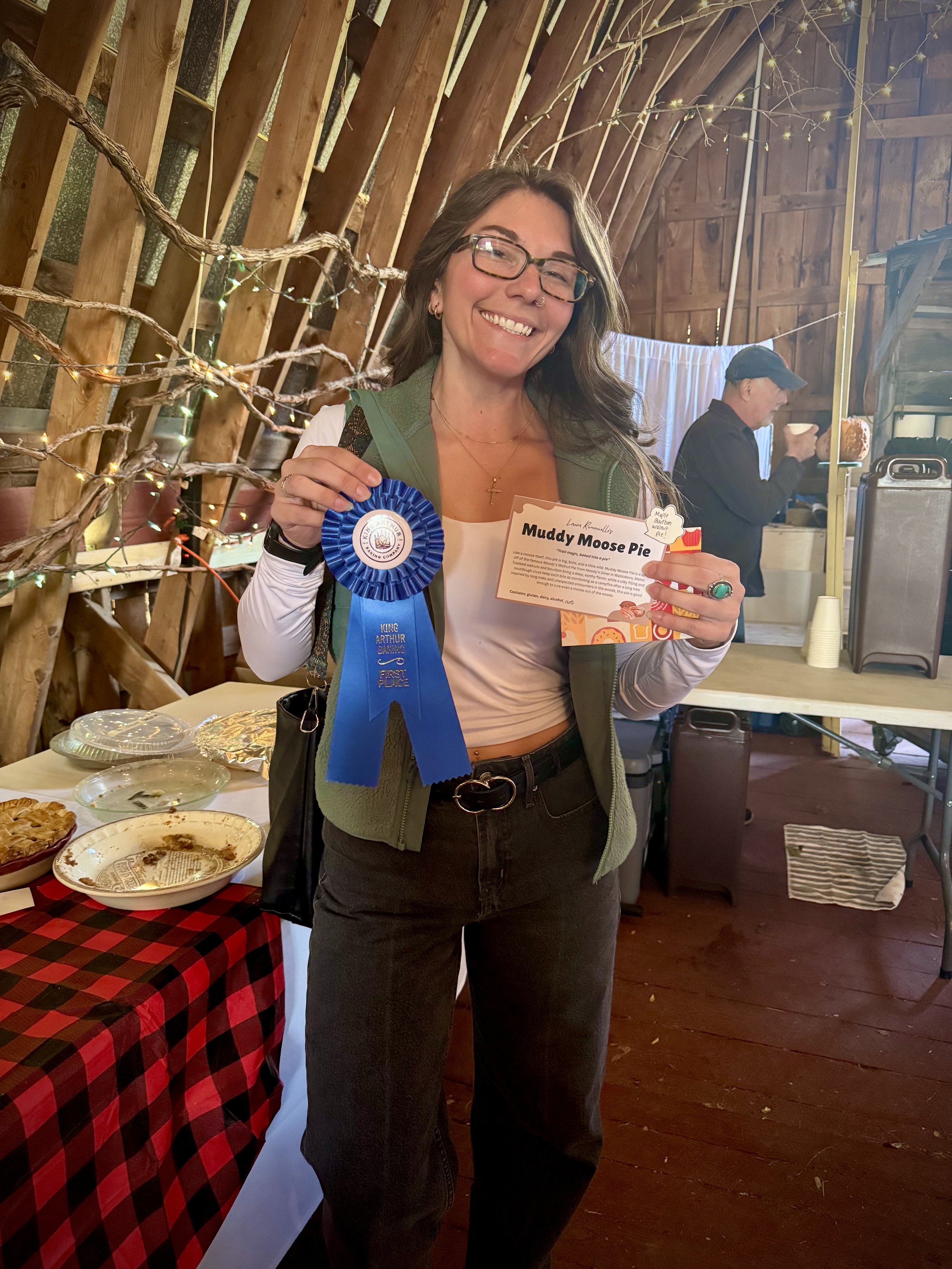Smiling woman holding a blue ribbon and a certificate at an indoor event with a rustic wooden background and a table with pie dishes. She just won first prize at the Southern Vermont Flannel Festival Bake Off!