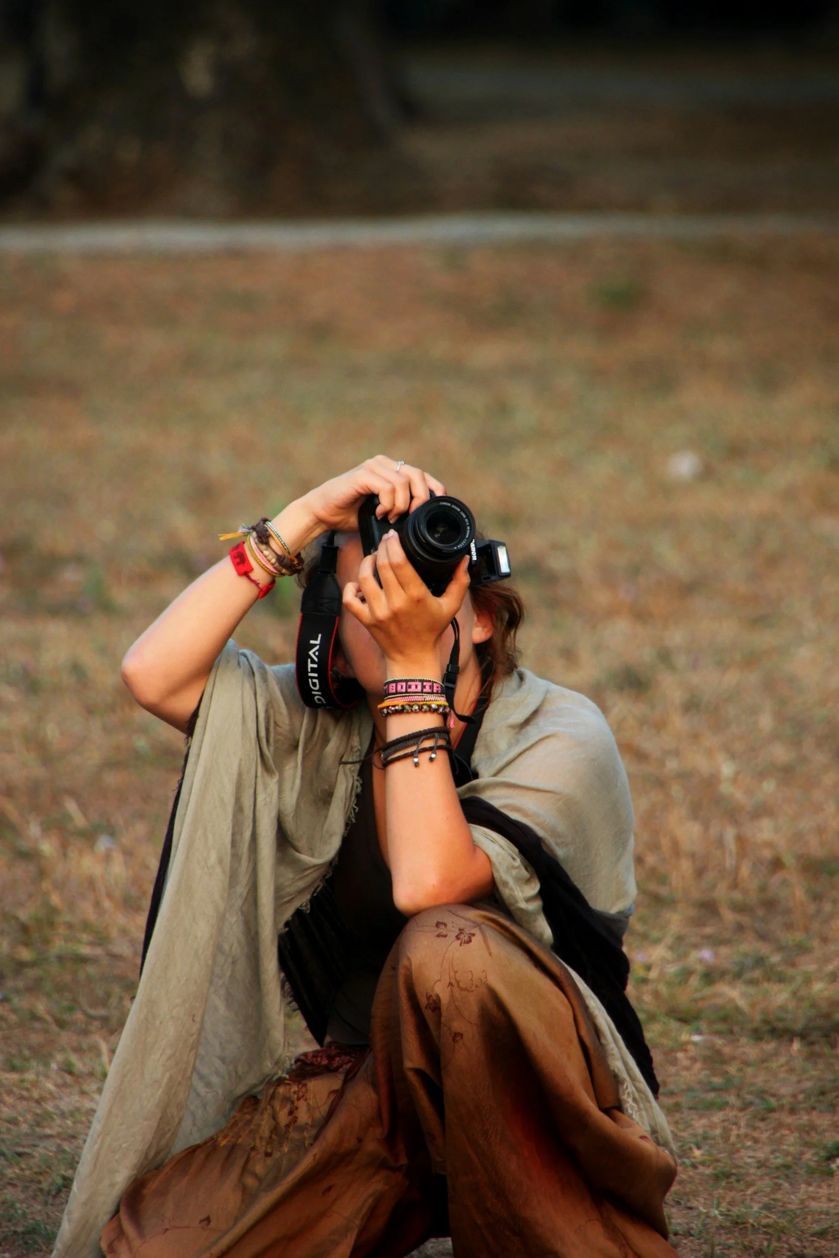 A person sitting outdoors on grass, holding a camera up to their face, taking a photograph. They are wearing multiple bracelets, a beige shawl, and loose brown pants.