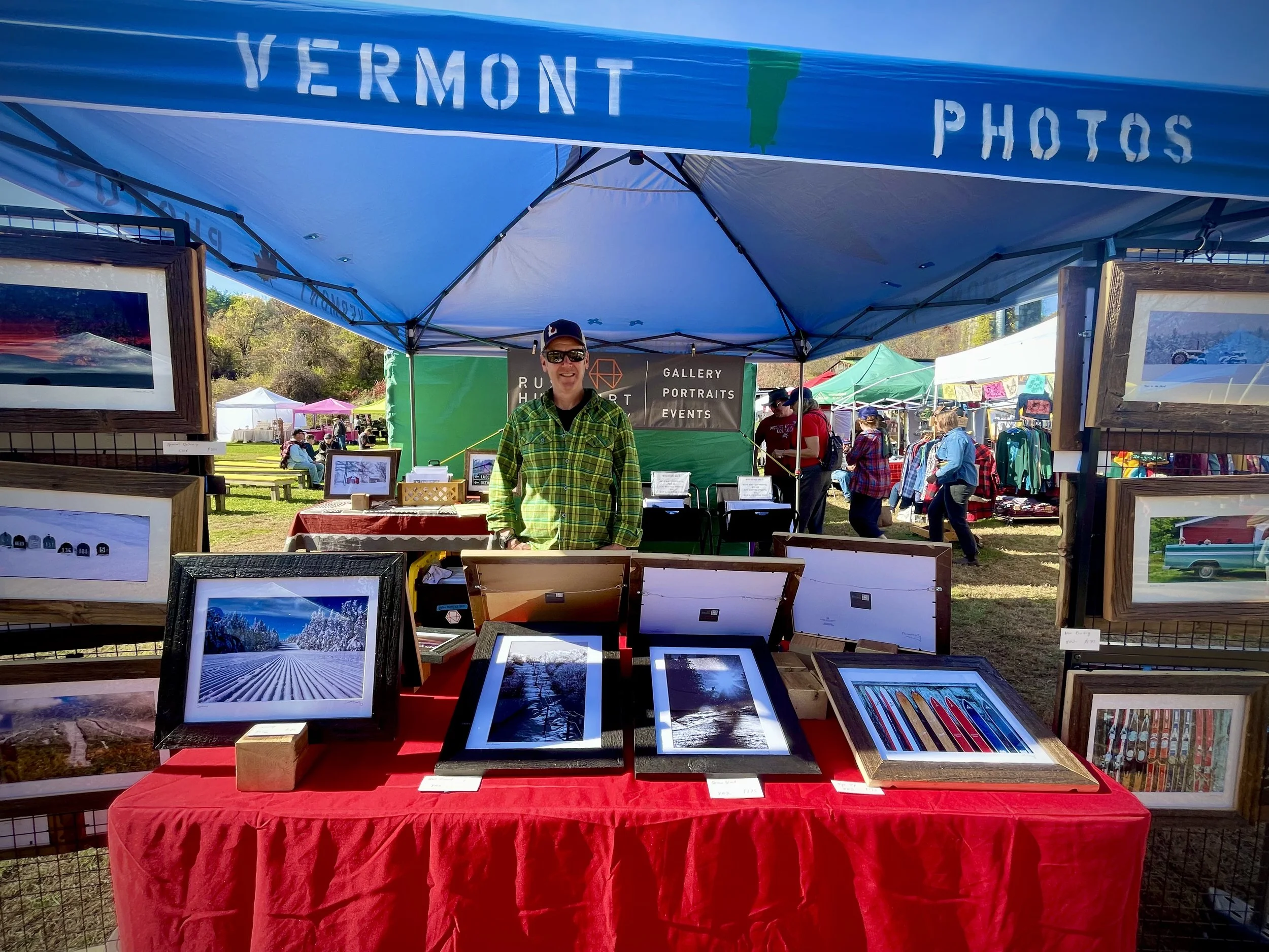 Photographer at an outdoor art market booth displaying framed landscape photographs, with other vendor tents and shoppers in the background.