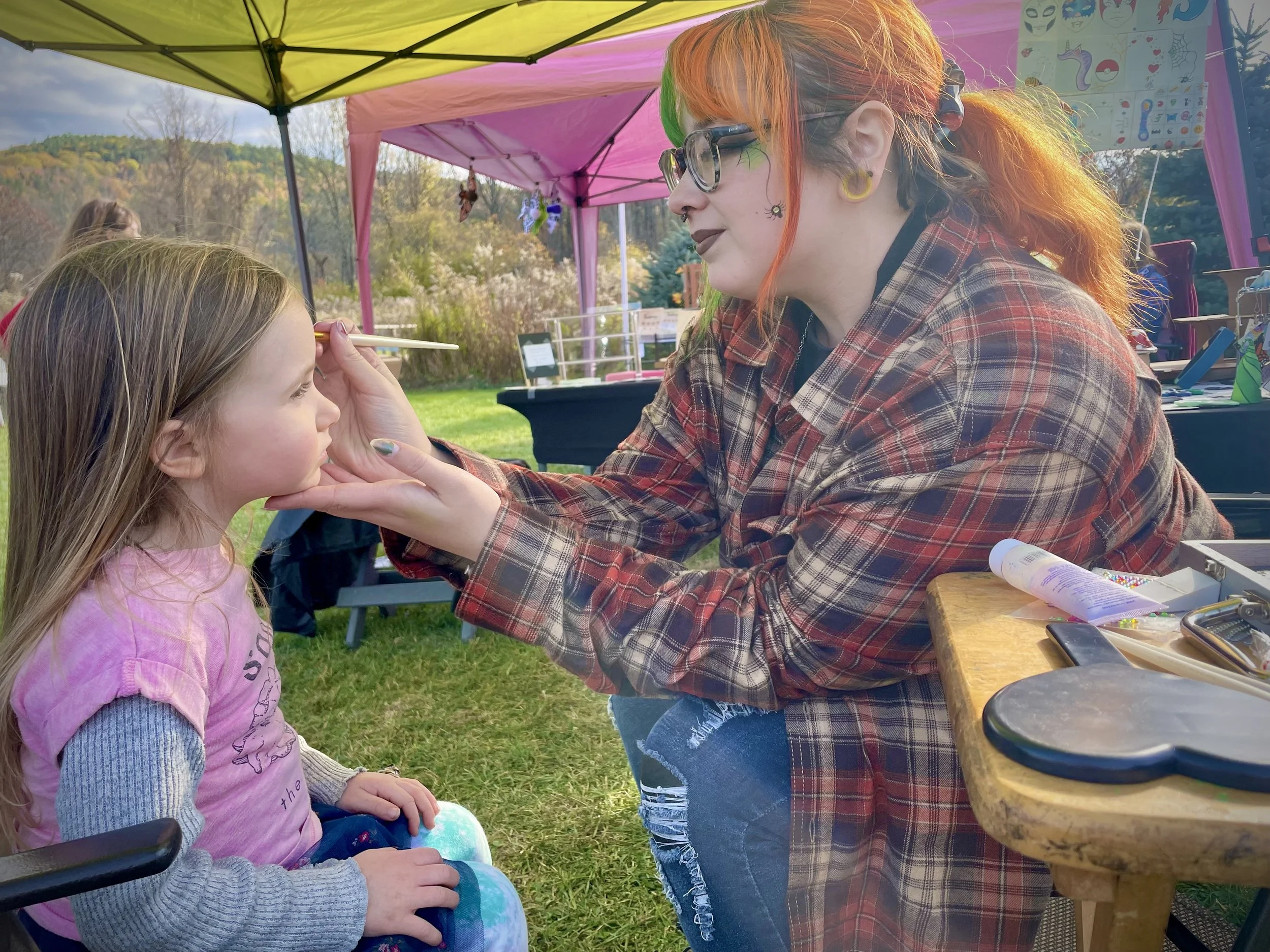 A woman with colorful hair and glasses is painting a design on a young girl's face at an outdoor festival with pink and yellow tents in the background.